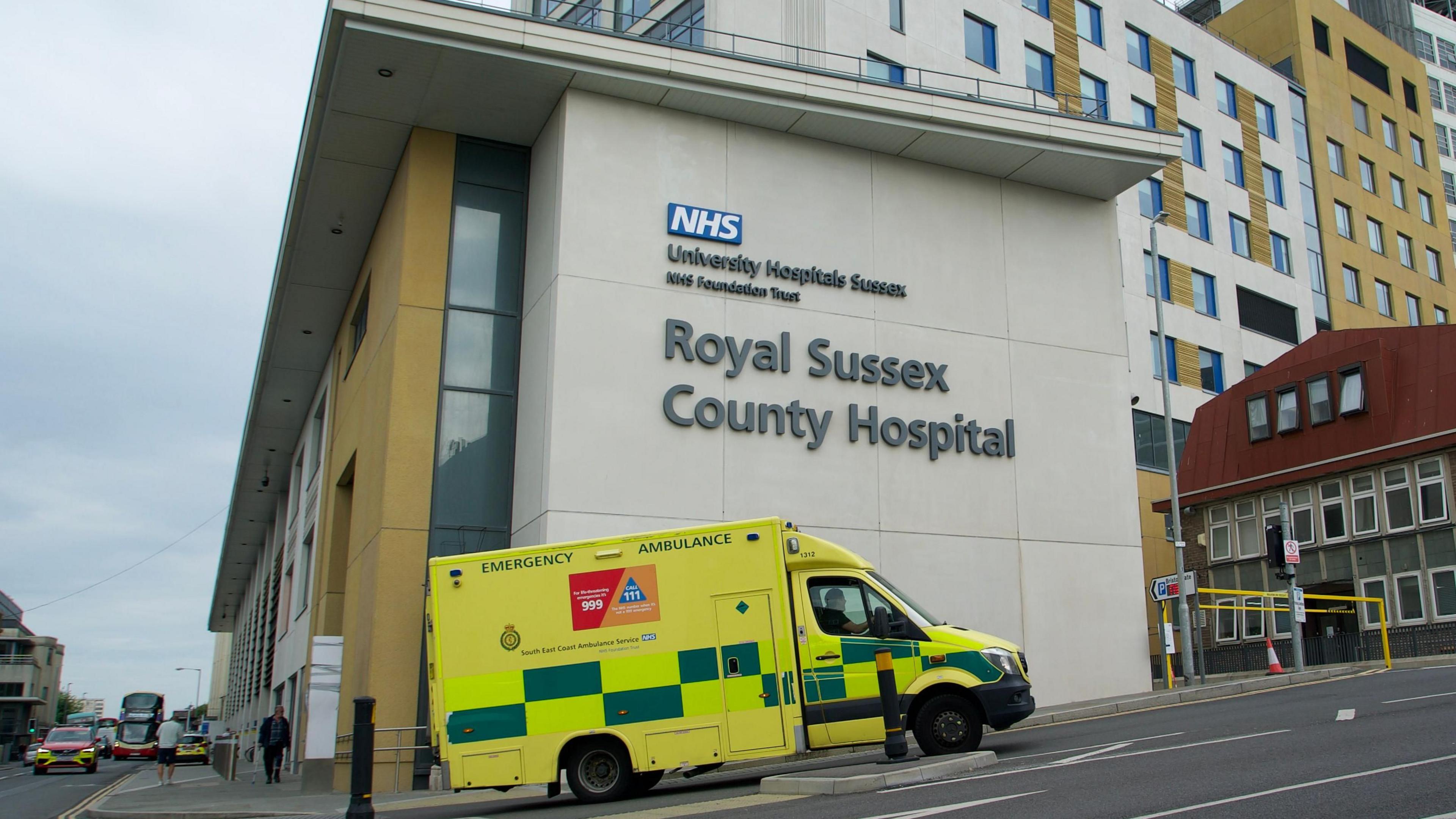 Side on shot of the Royal Sussex County Hospital in Brighton with a yellow ambulance driving up the road beside it.