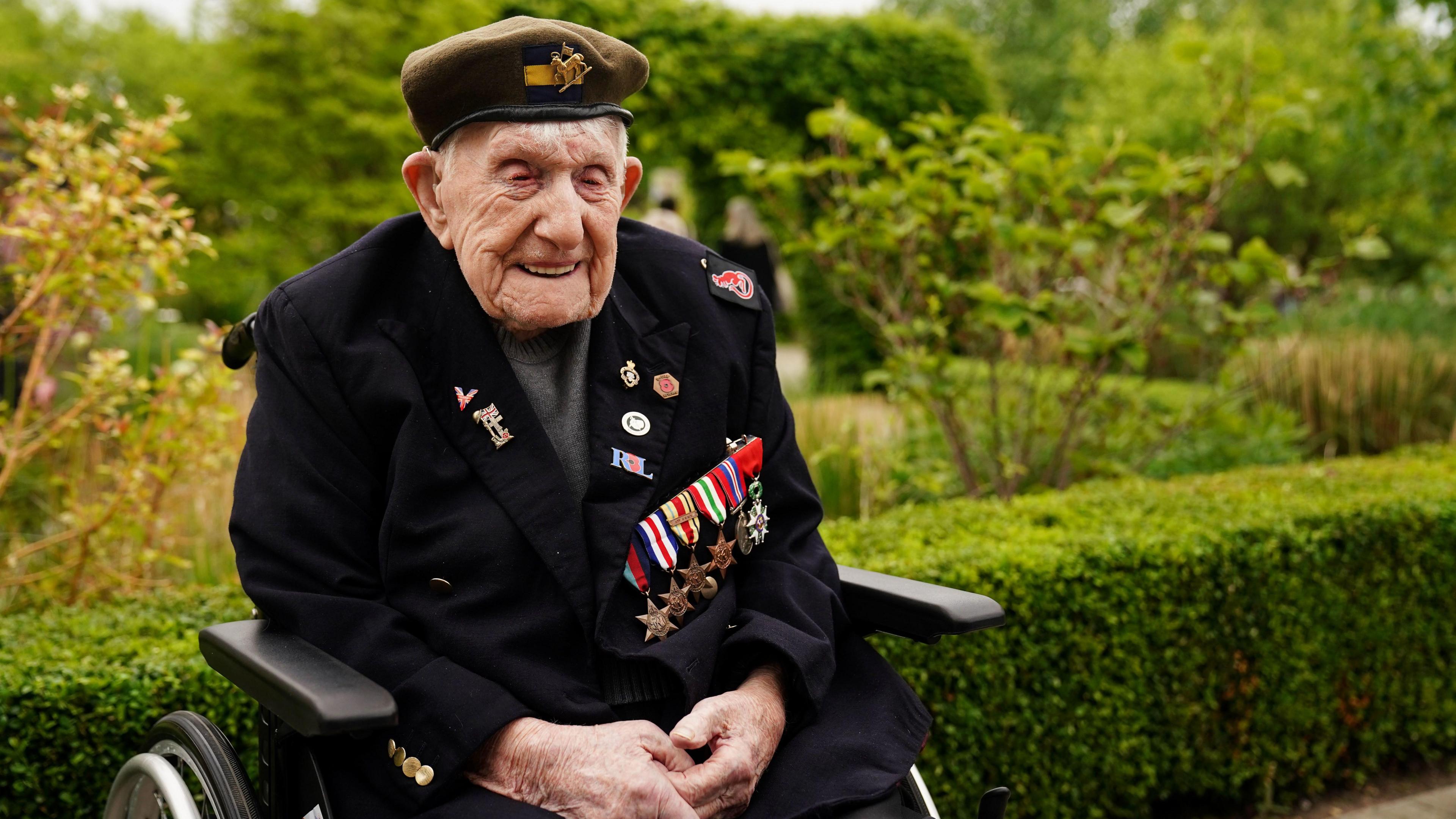 A veteran wearing a beret and a number of medals on a blue jacket, sits smiling in a wheelchair.