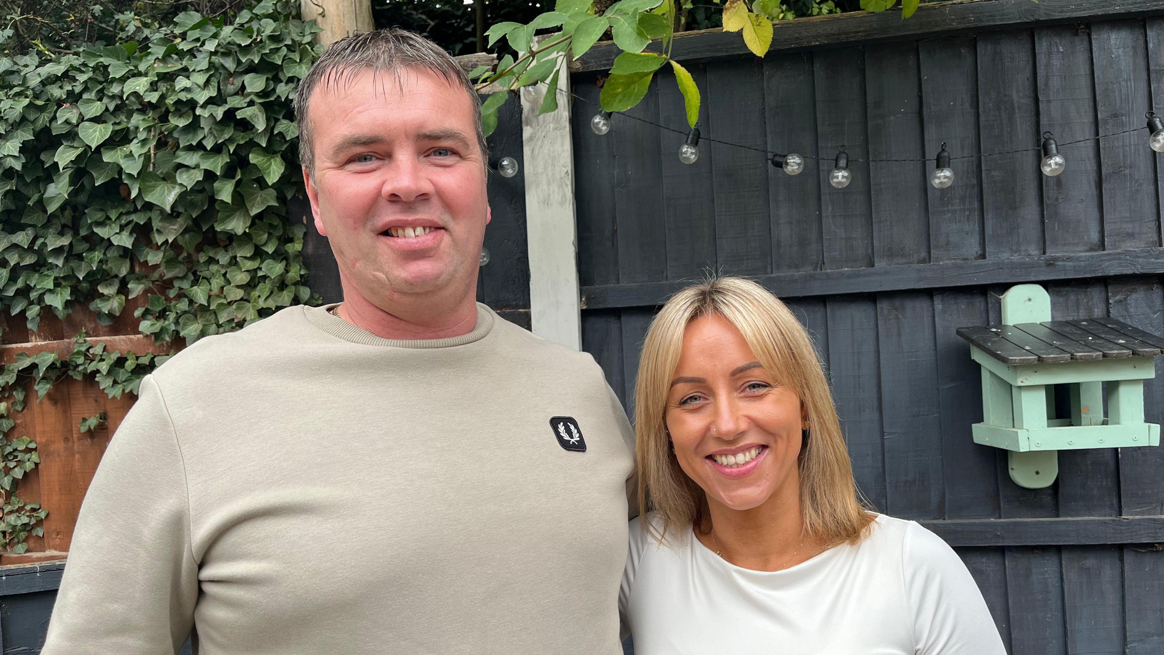 Kevin and Jodie stood side by side with their arms around each other and smiling. Kevin is wearing a beige jumper, and Jodie is wearing a white long sleeved top. The couple are stood in their garden, with a grey wooden fence behind them that's got fairy lights strung along the top of it.