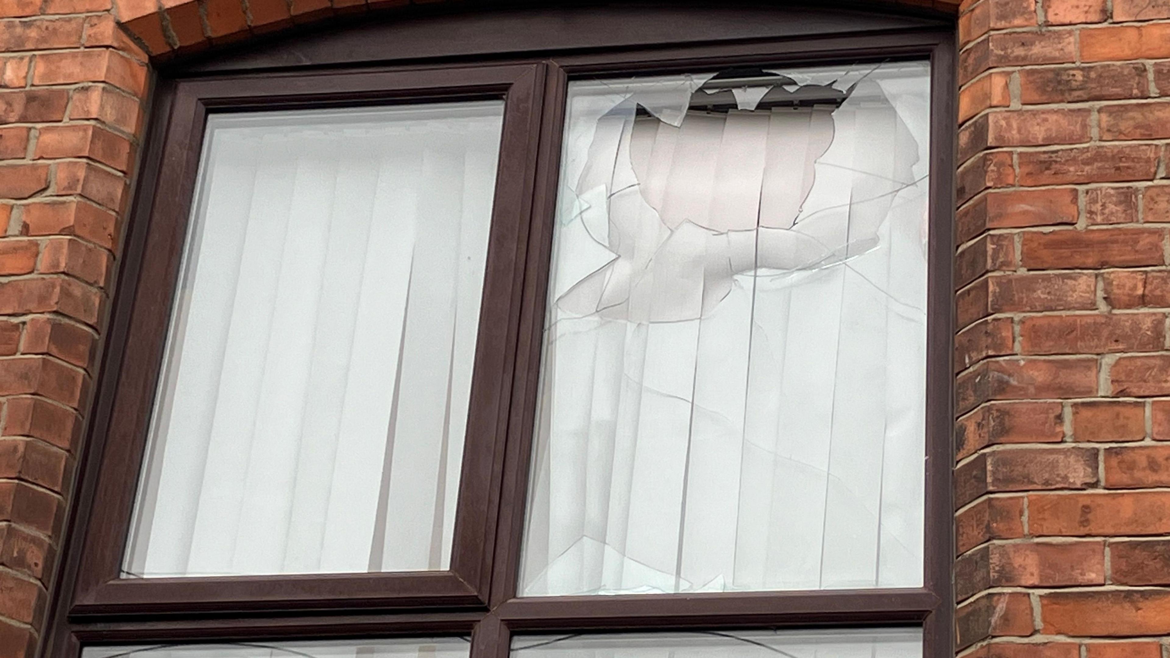 A window smashed through with blinds behind - the house is red brick with brown window panes. 