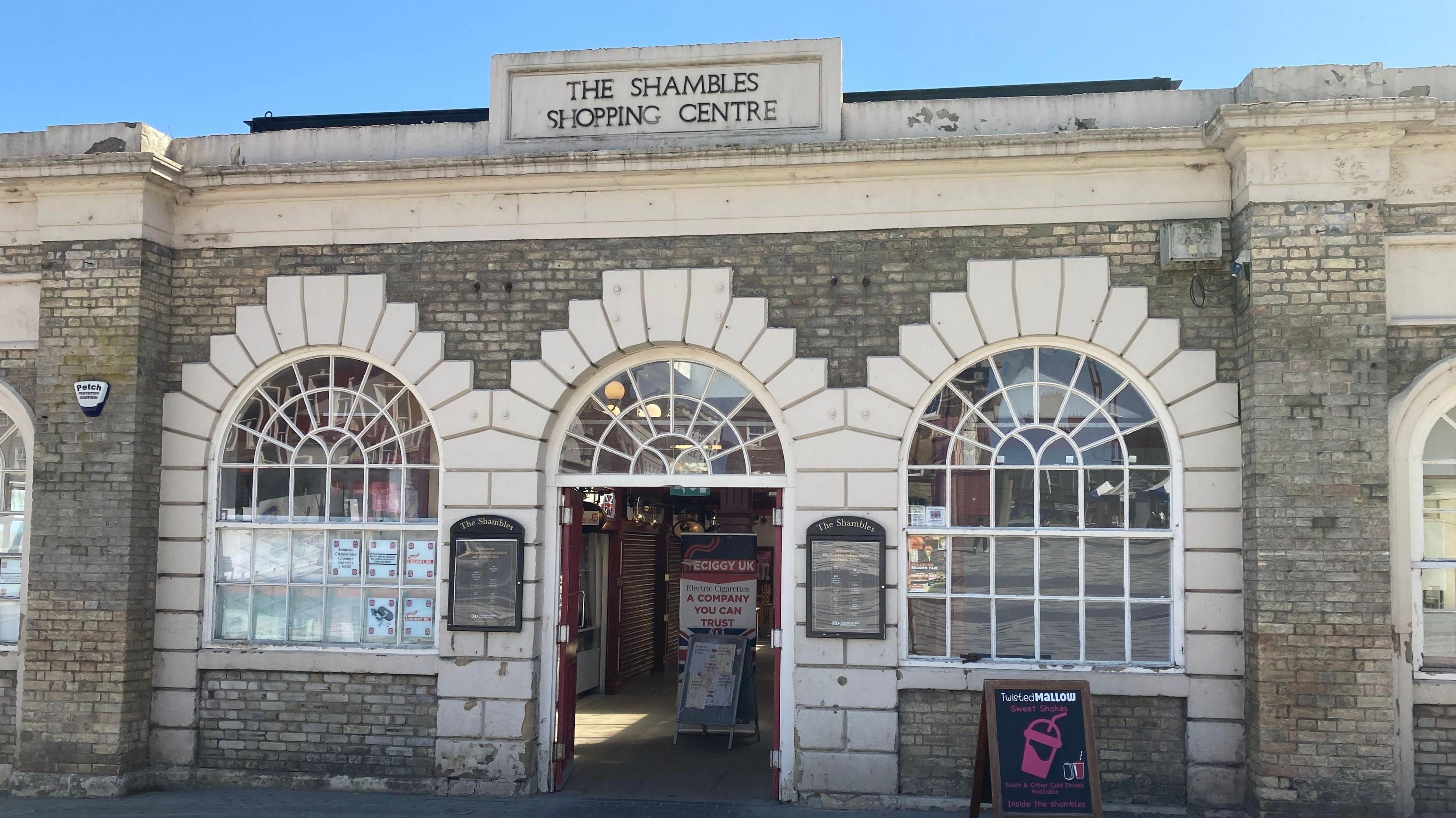 The entrance to 'The Shambles Shopping Centre' with a sign outside for the Twisted Mallow sweet shop. Three white stone arches are on the front of the building.