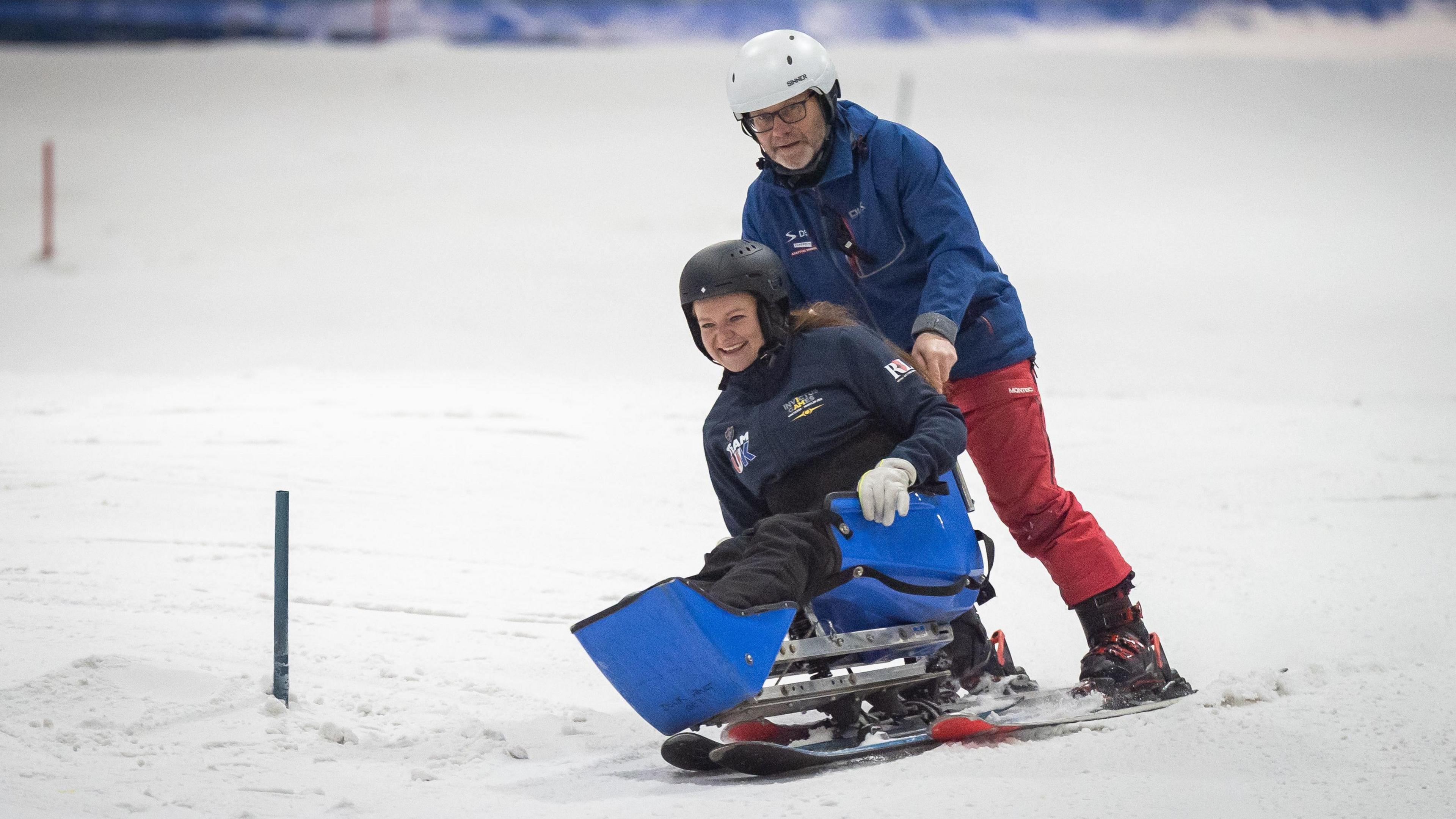 A woman is wearing a helmet while using a set of sit skis, a kind of sled on skis. Behind her is a man also wearing a helmet and skis. His hand is holding the seat as they make their way down a ski slope.