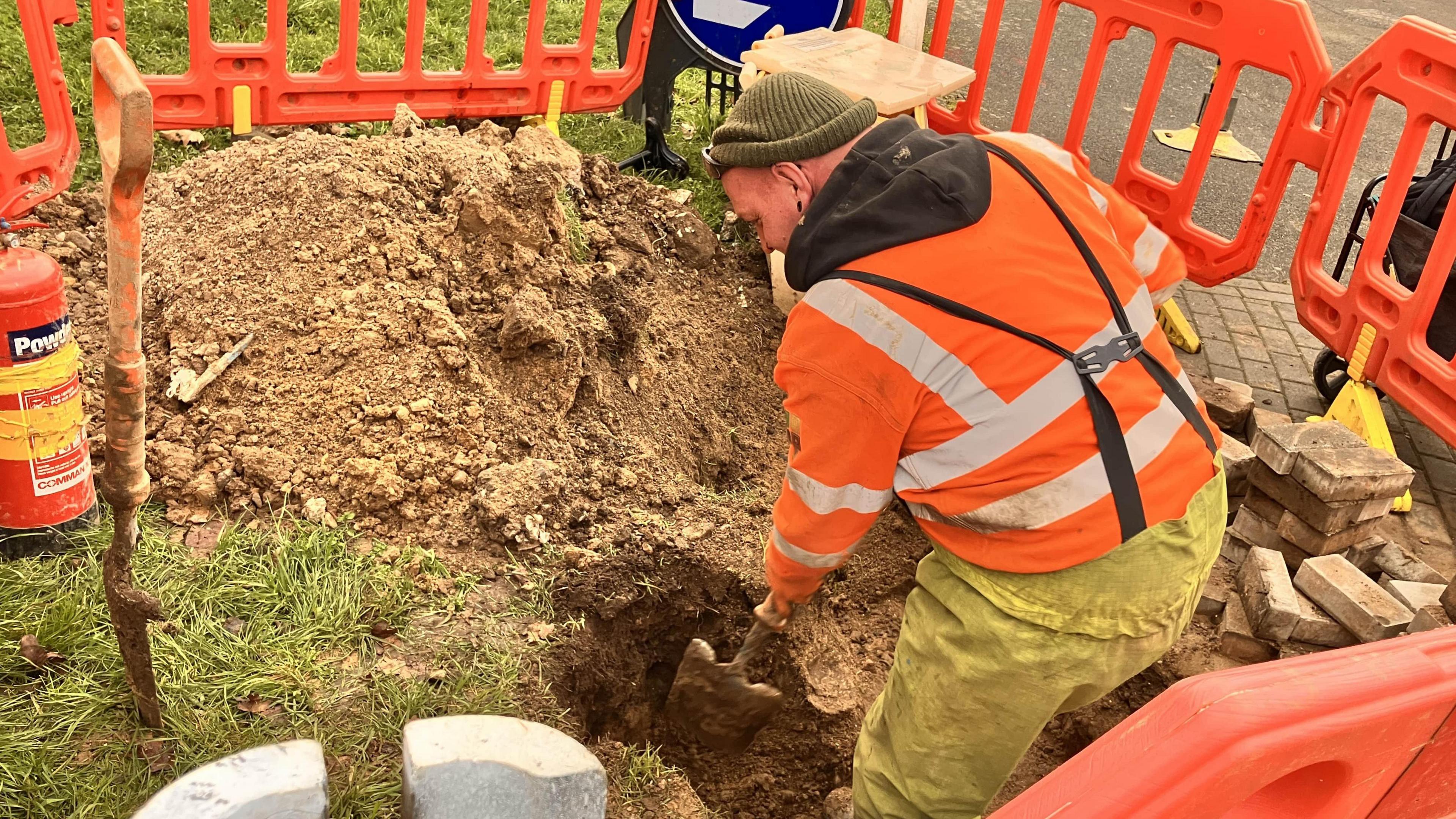 An engineer digs a hole in the ground with a shovel. He wears an orange hi-vis jumper, knitted hat and trousers. Orange temporary fencing surrounds him.