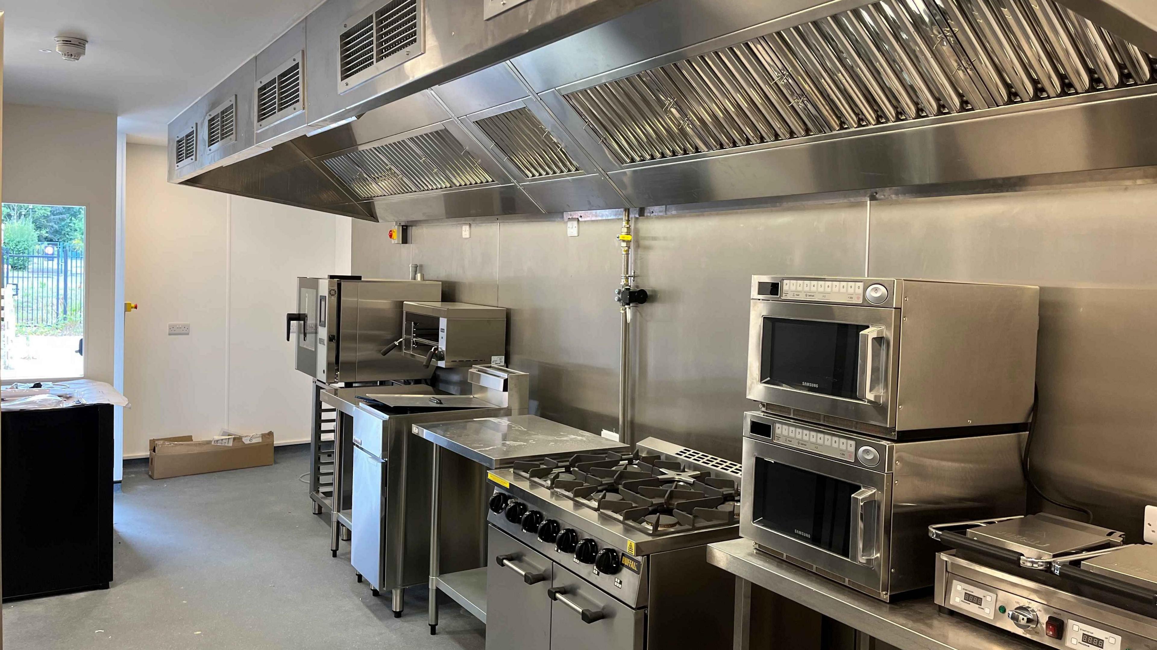 An industrial kitchen with stainless steel catering equipment lined up under a large extractor fan. 