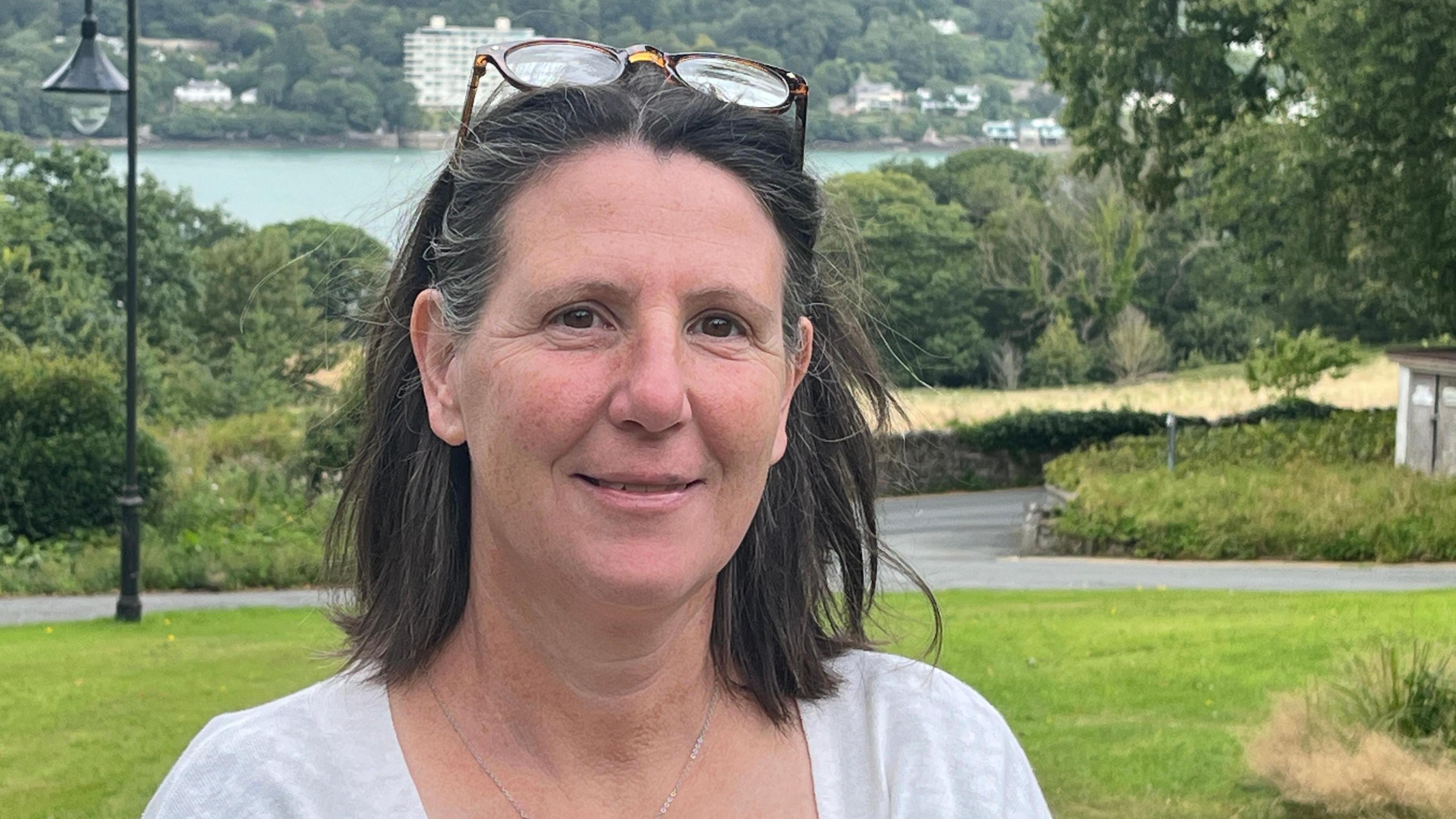 Llun pen ac ysgwyddau o Dr Linda Osti, dynes gyda gwallt brown at ei hysgwyddau, yn sefyll mewn man gwyrdd gyda'r Fenai a rhan o arfordir Ynys Môn yn y pellter tu ôl iddi