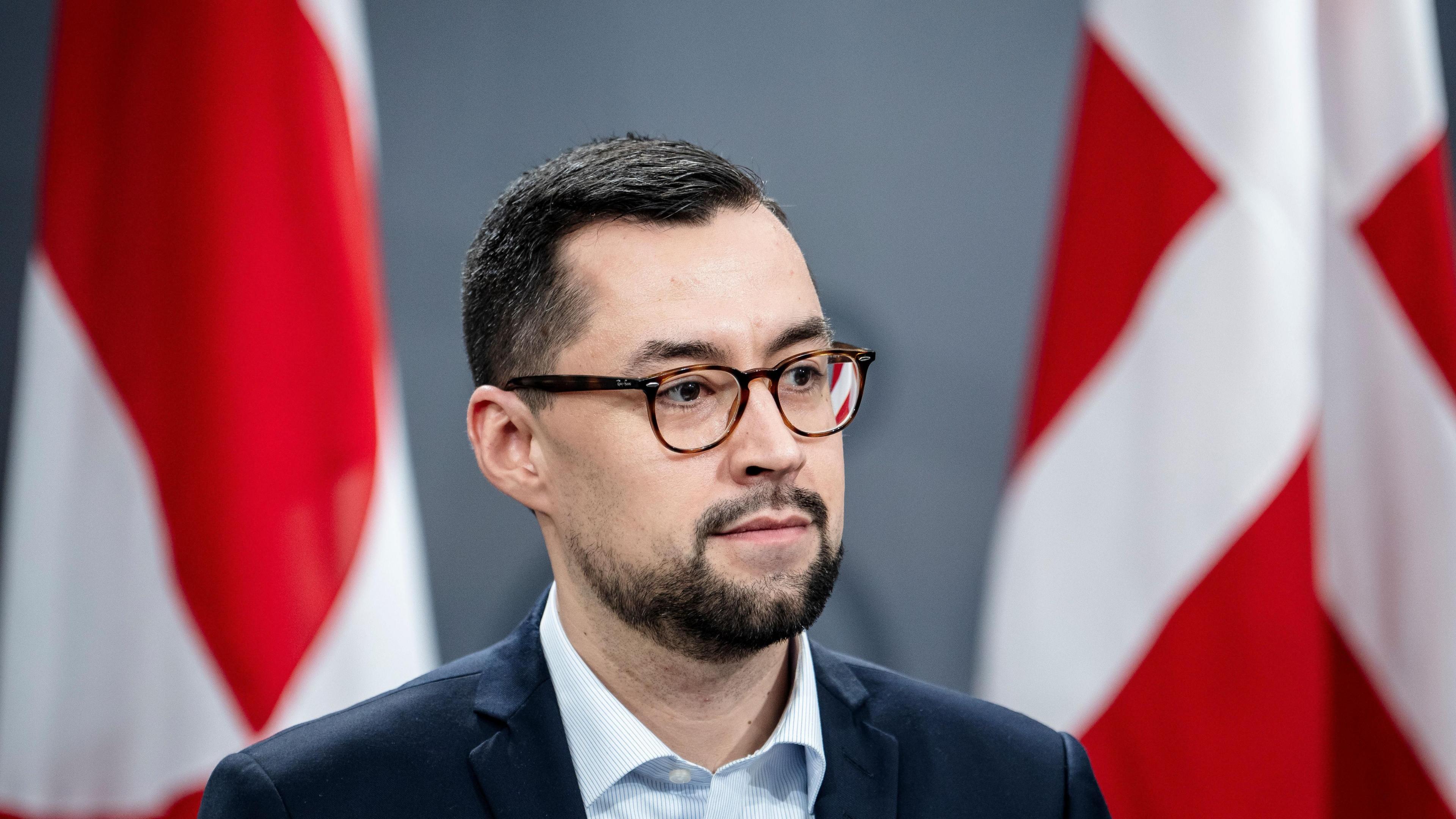  Mute Egede, a man with brown glasses and black hair and facial hair, stands in front of two Danish flags.