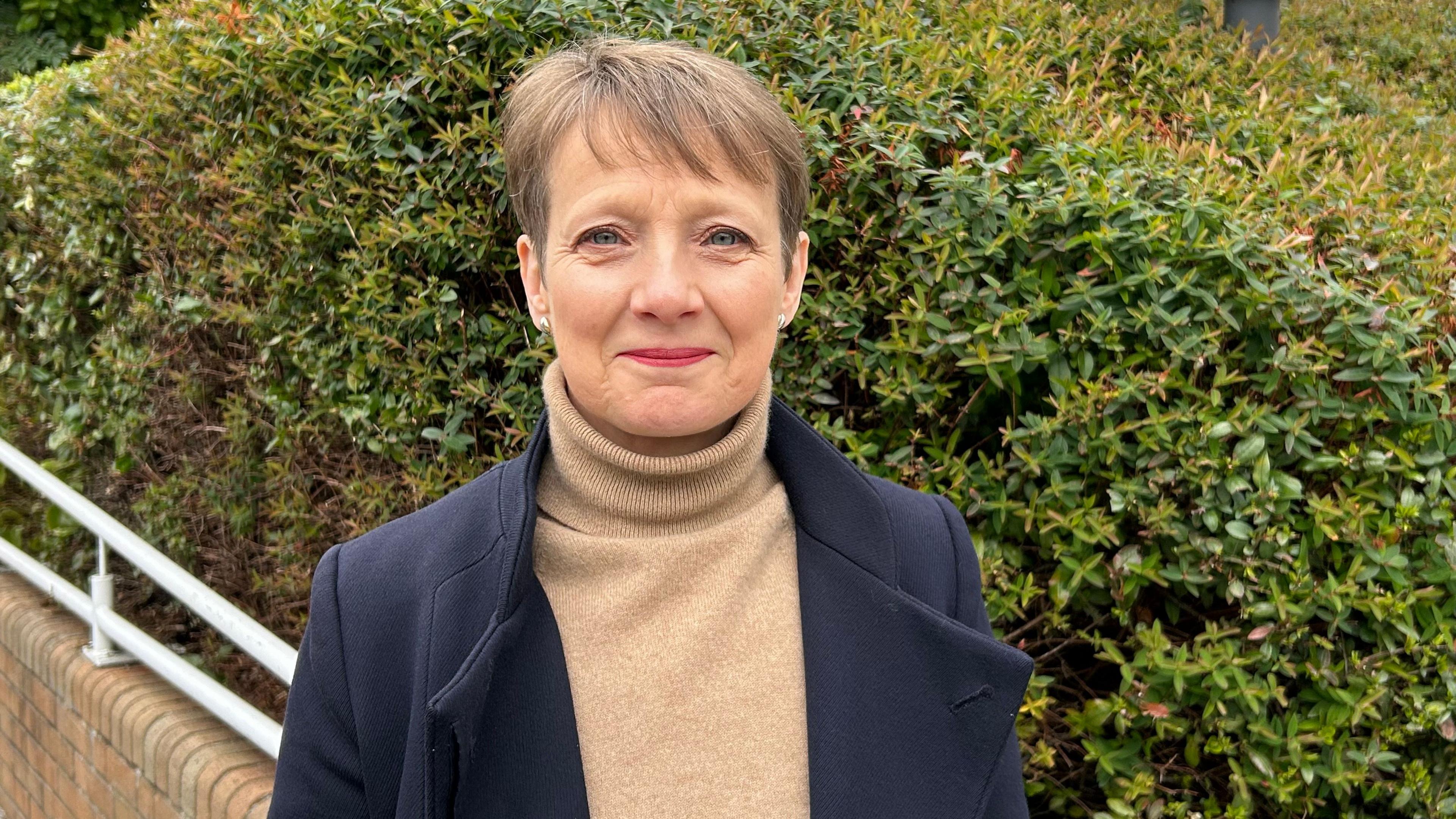 Clare wearing a beige turtleneck and dark jacket standing outside smiling at the camera. She has short brown hair, blue eyes and studded earrings.