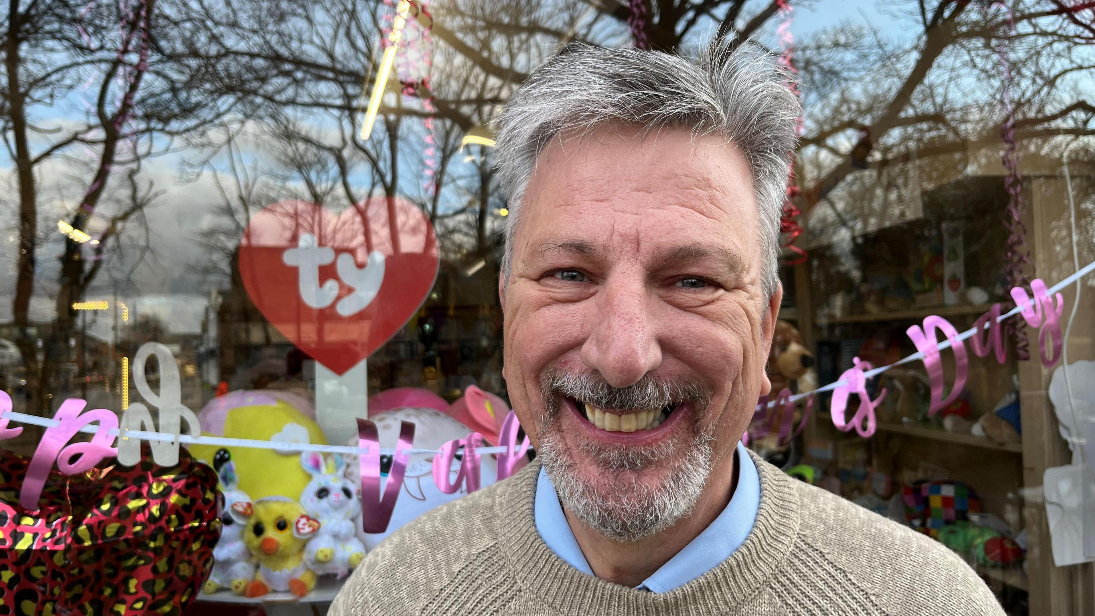 Nigel Lambert has grey hair sticking up and a grey beard and moustache. He is wearing a beige woollen jumper and a blue shirt collar is visible. He is standing in front of the window of a shop that sells balloons, greetings cards and gifts. Trees are reflected in the window.