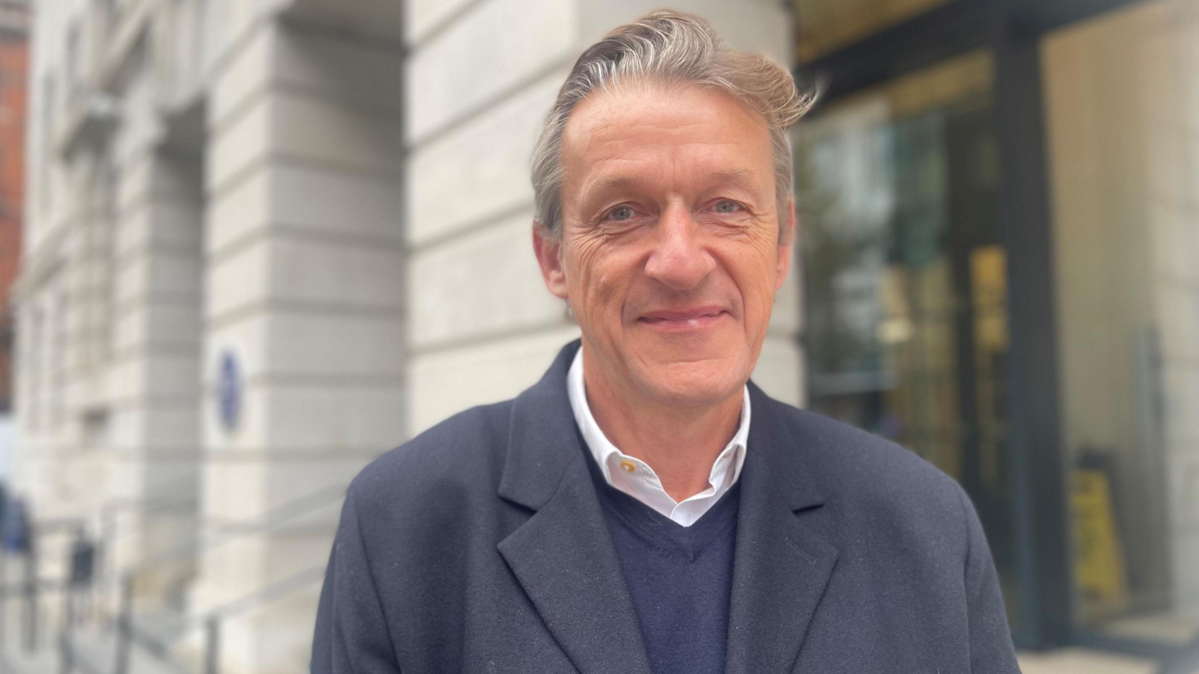 A man with greying blond hair and a blue trench coat stands outside a council building
