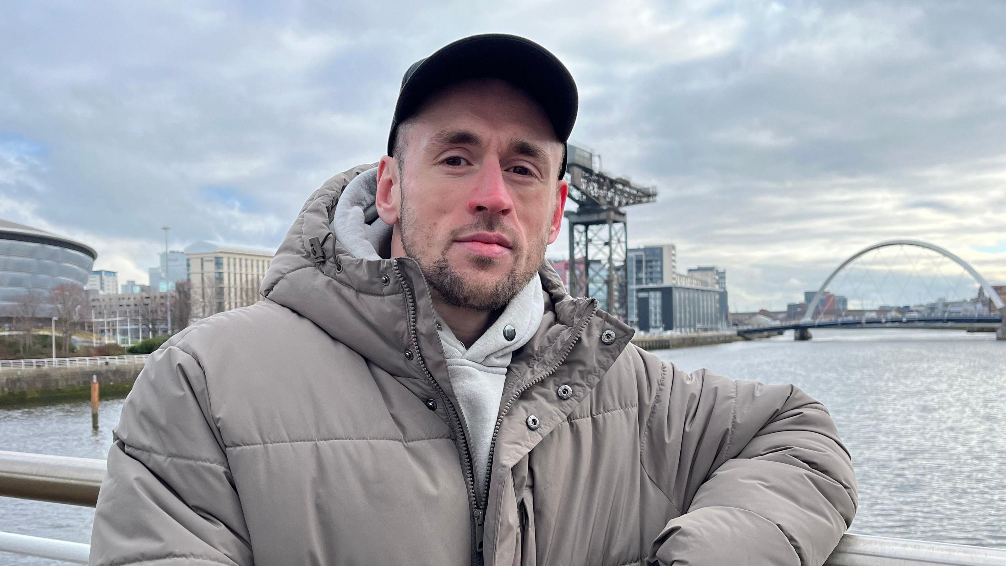 Jordan Robertson stands over a bridge on the River CLyde, looking at the camera with his arms crossed. He's wearing a puffy grey jacket and has a baseball cap on. 