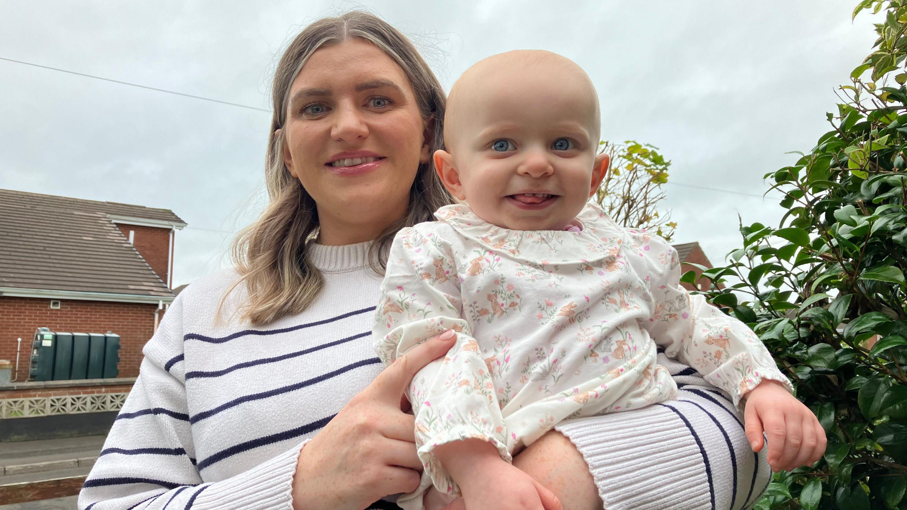 Lauren is holding Anna, they are both wearing white and both smiling into the camera. They are standing in a garden with a bush and a house in the background.