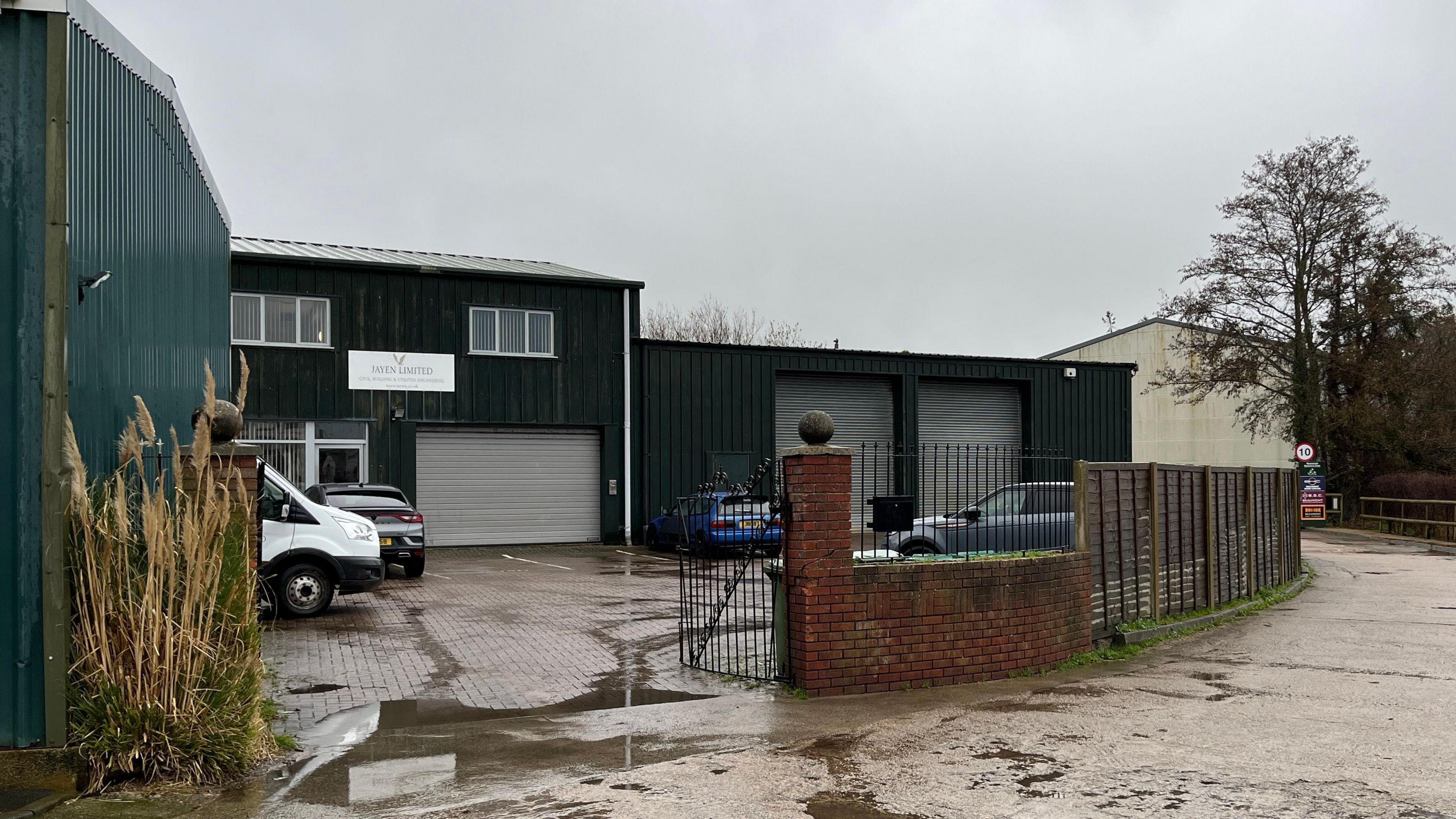 A green building in an L-shape with a car park in front within gates on a rainy day.