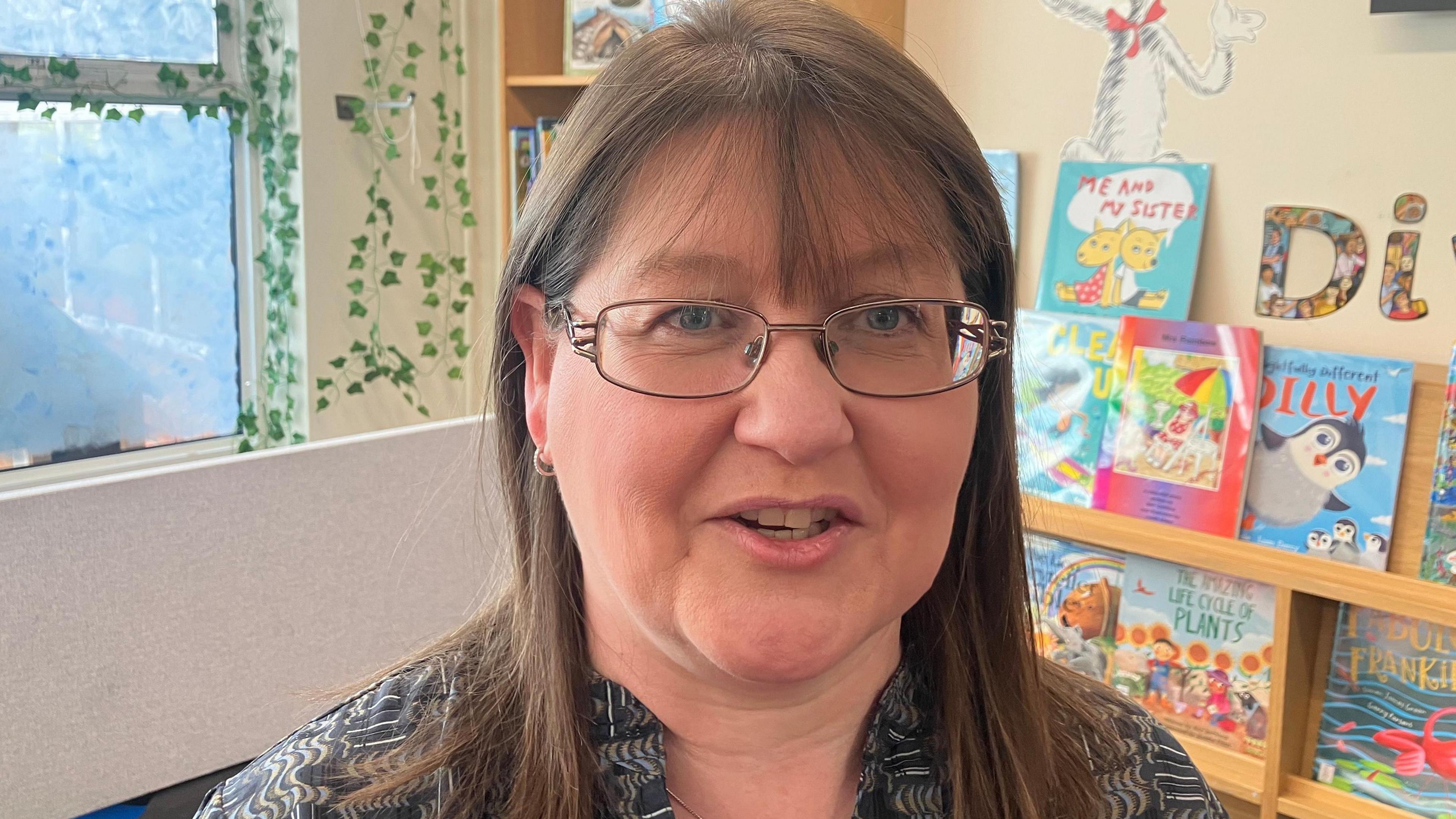 Teresa Devlin has long brown hair and wears glasses. Behind her is a wall with a painting of leaves. There are shelves with books on them.