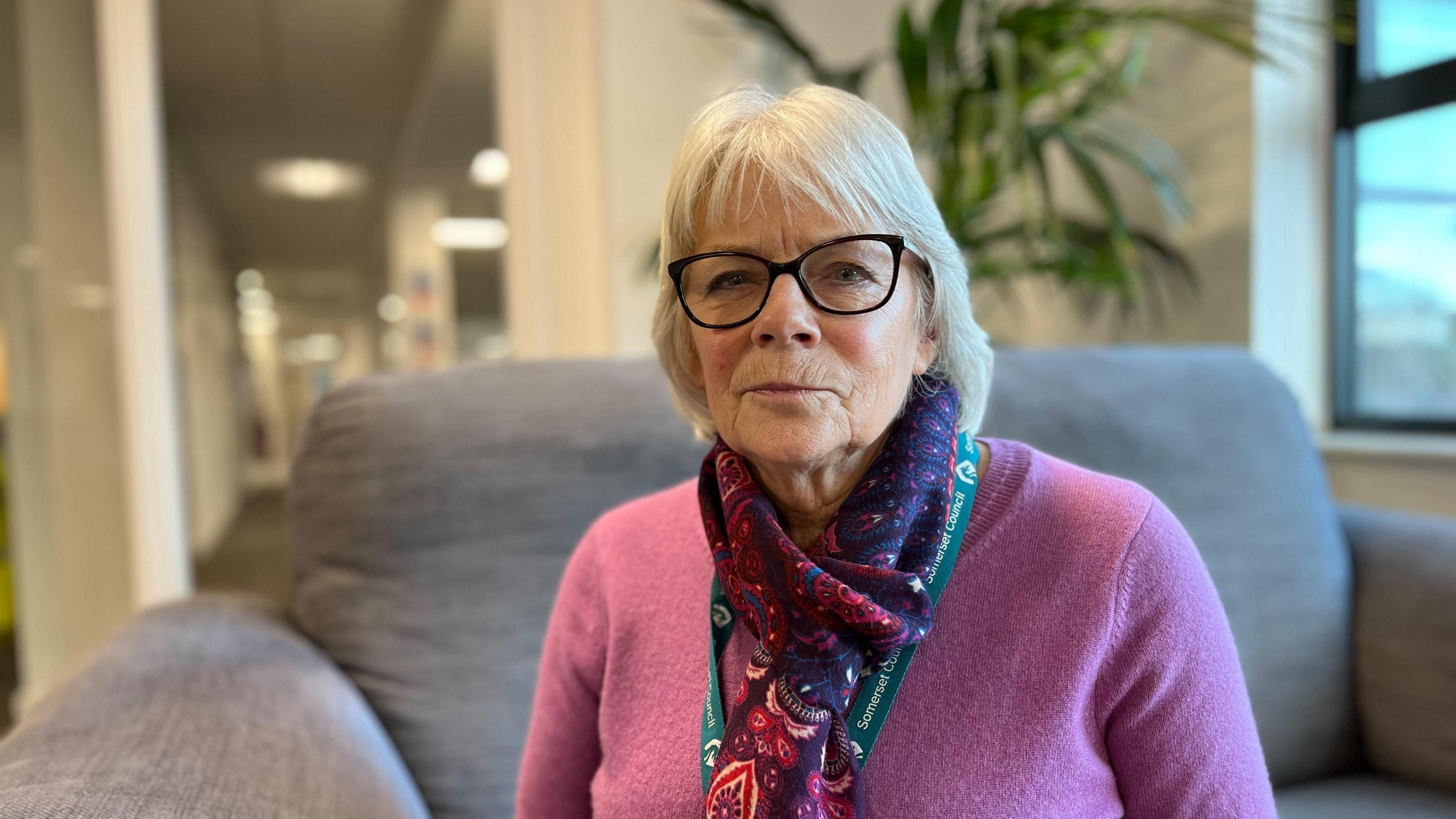 A woman with black-rimmed glasses, in a purple cardigan and colourful scarf sitting on a grey sofa in an office in front of a pot plant.