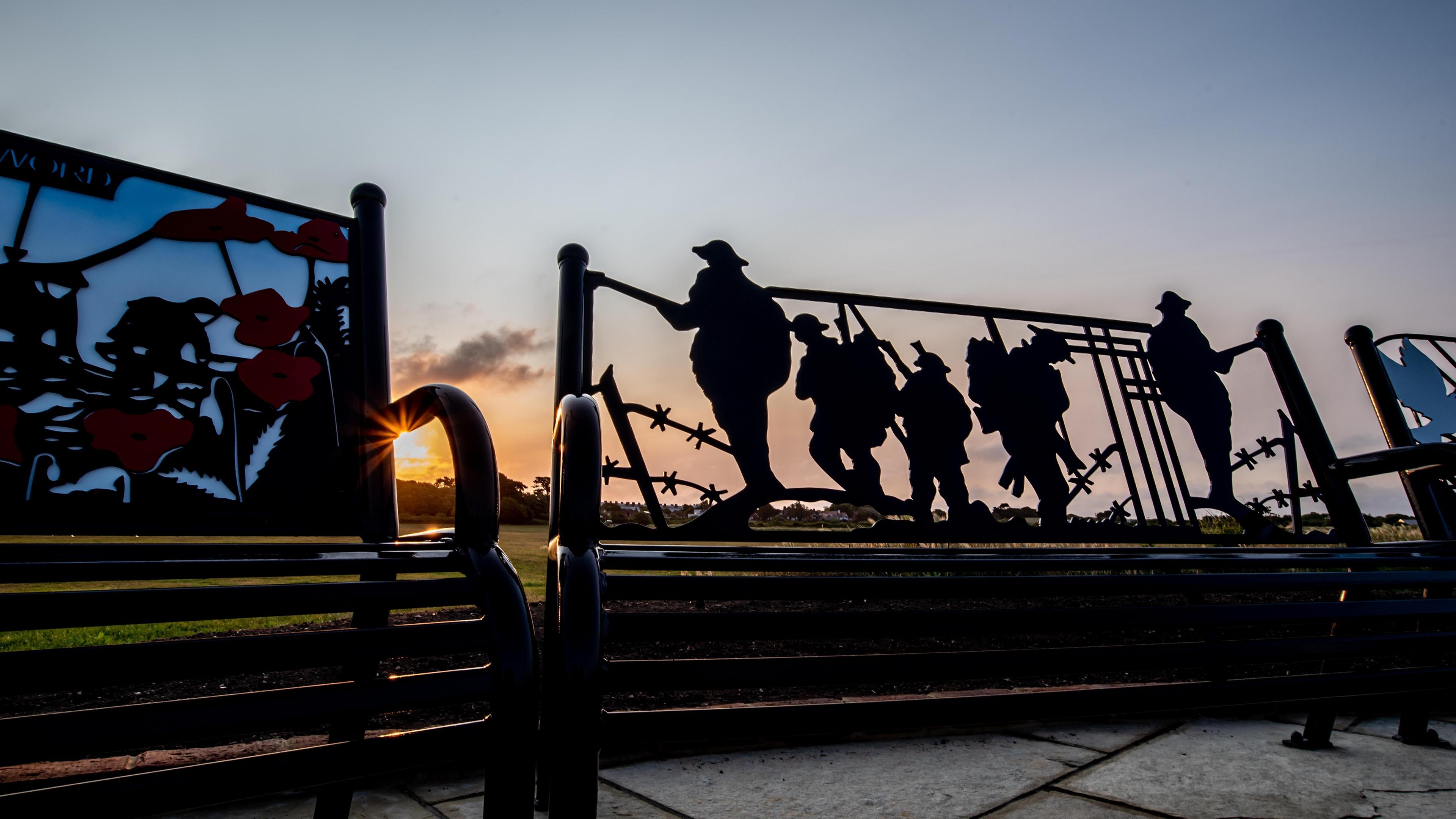 Silhouettes of soldiers and barbed wire on the back of bench. You can see the sun setting behind and an orange sky. There are small clouds in the sky. One of the three benches has poppy sculptures on the back.