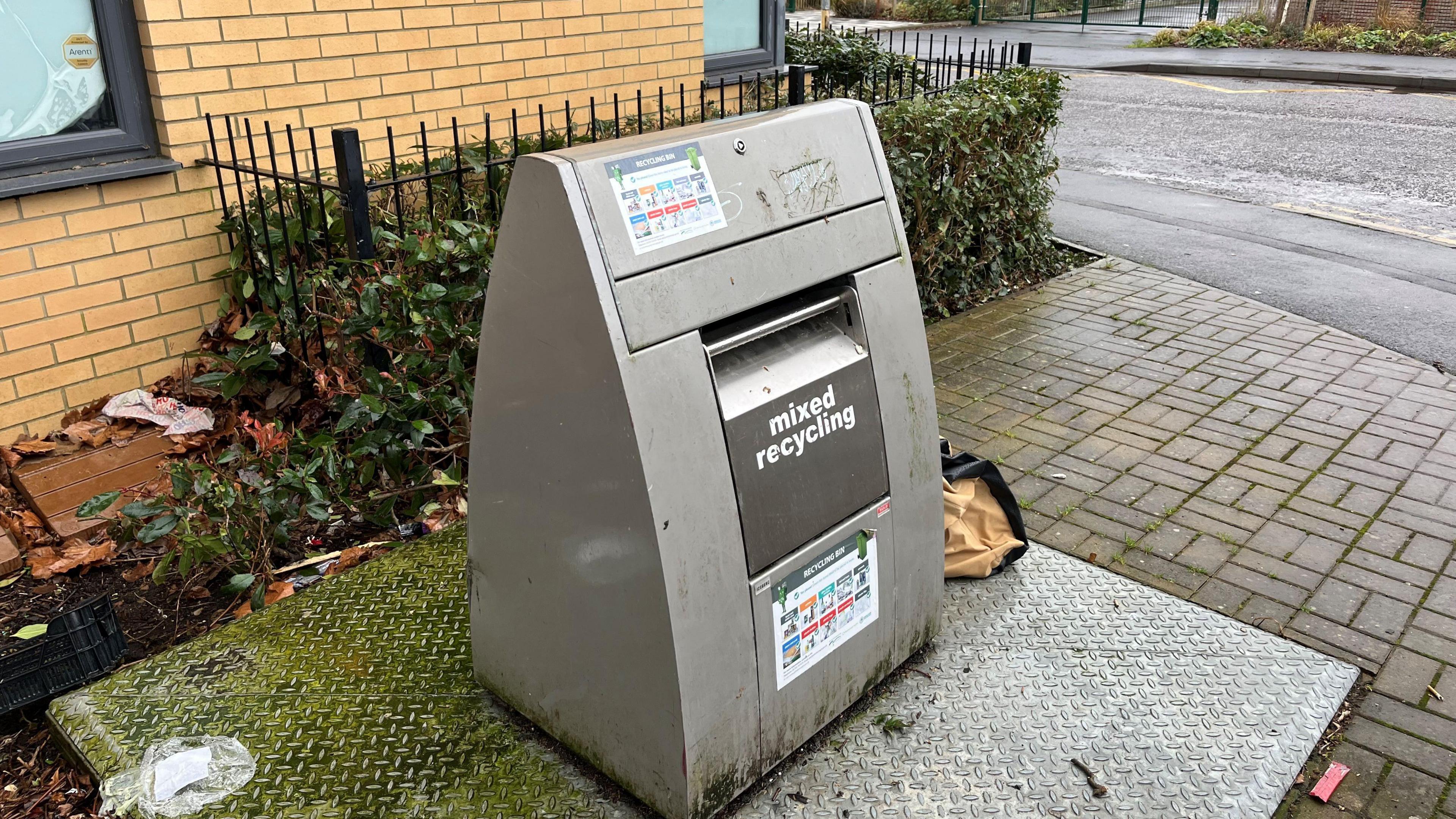 A triangular shaped silver coloured metal contraption which is a chute to deposit rubbish in, there are items of rubbish around it and on the block paved area adjacent to the tarmac footpath. Behind is the corner of one of the rooms in the block of flats.