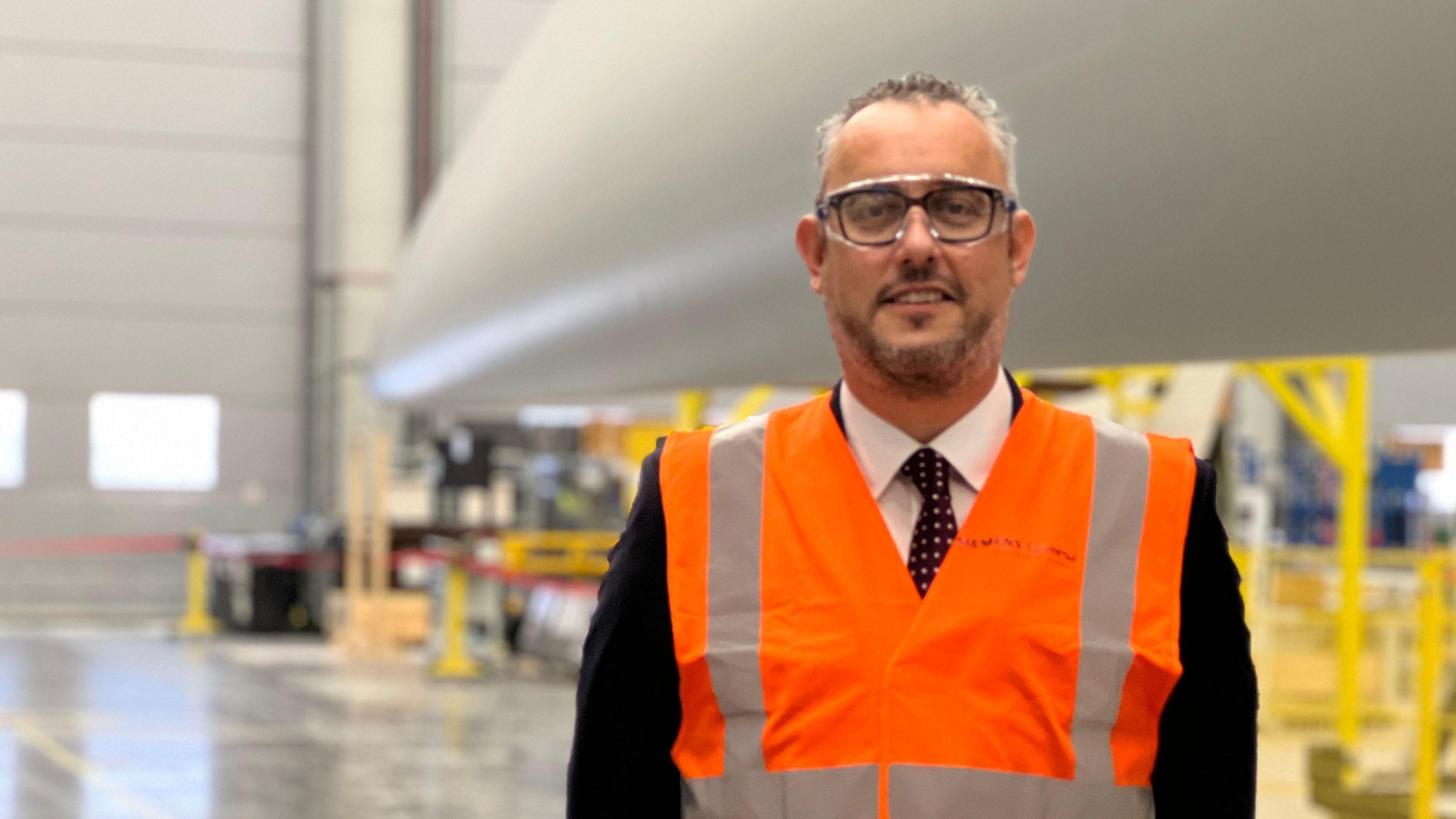 Darren Davidson stands in front of a wind turbine blade within Siemens Gamesa's Hull manufacturing plant. He has short grey hair and a short dark beard, and wears glasses, a black suit and an orange hi-vis vest.