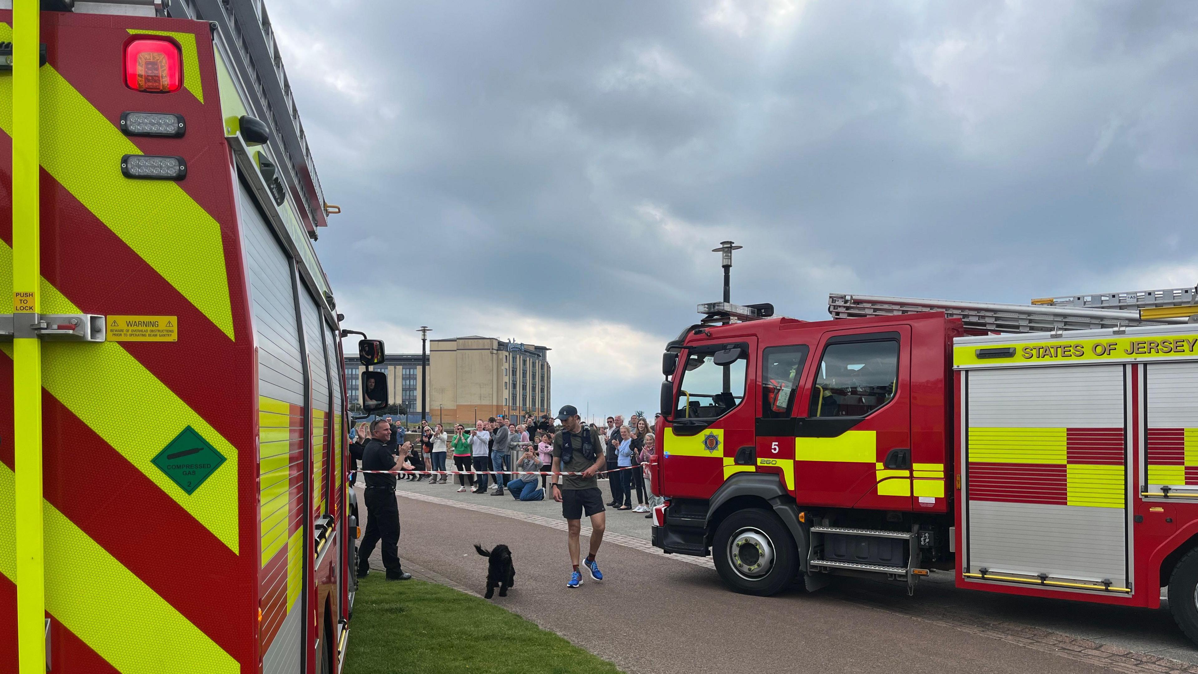 Matt gets to the finish line which has been held up by two fire engines as a crowd cheers him on and his black dog is by his side after the run.