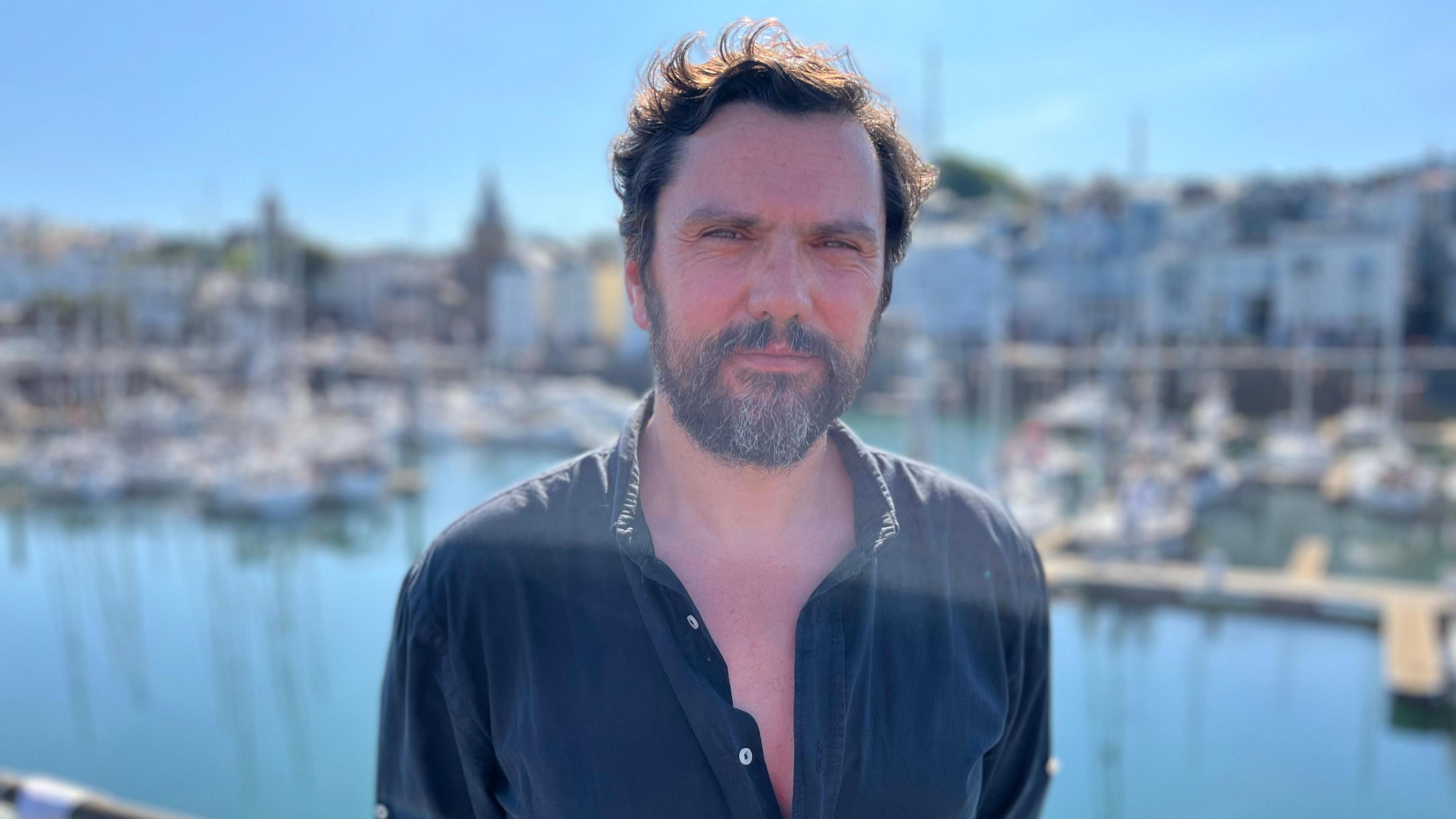 Oliver is wearing a blue shirt and has short dark brown hair as well as a beard. He is standing at the harbour in St Peter Port in Guernsey.