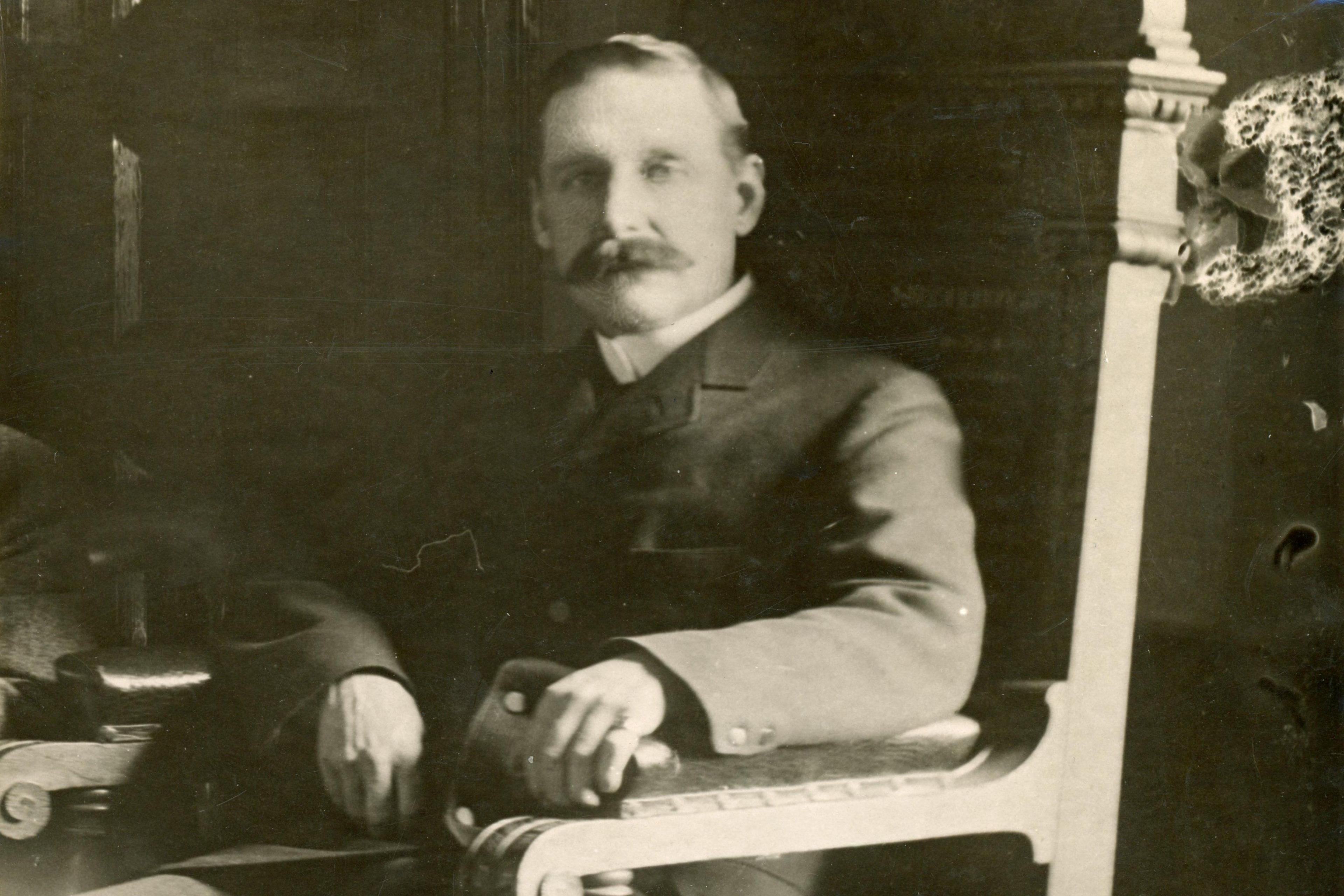 John is sitting in a grand wooden chair. He has a moustache and is looking at the camera. It is a black and white photograph. He is wearing a dark suit jacket and white collar.