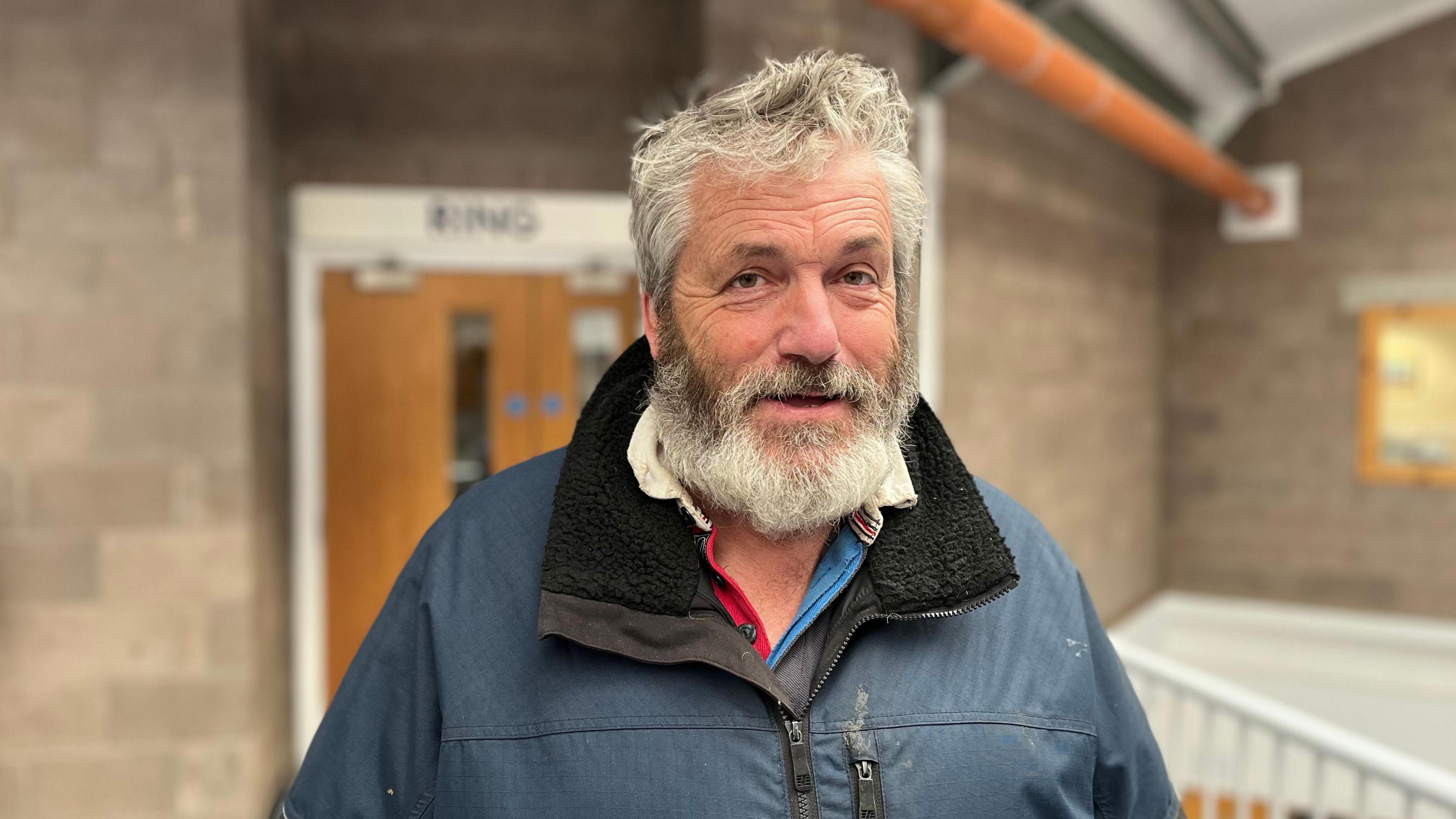 A grey haired man with a grey beard looks at the camera with a door behind him