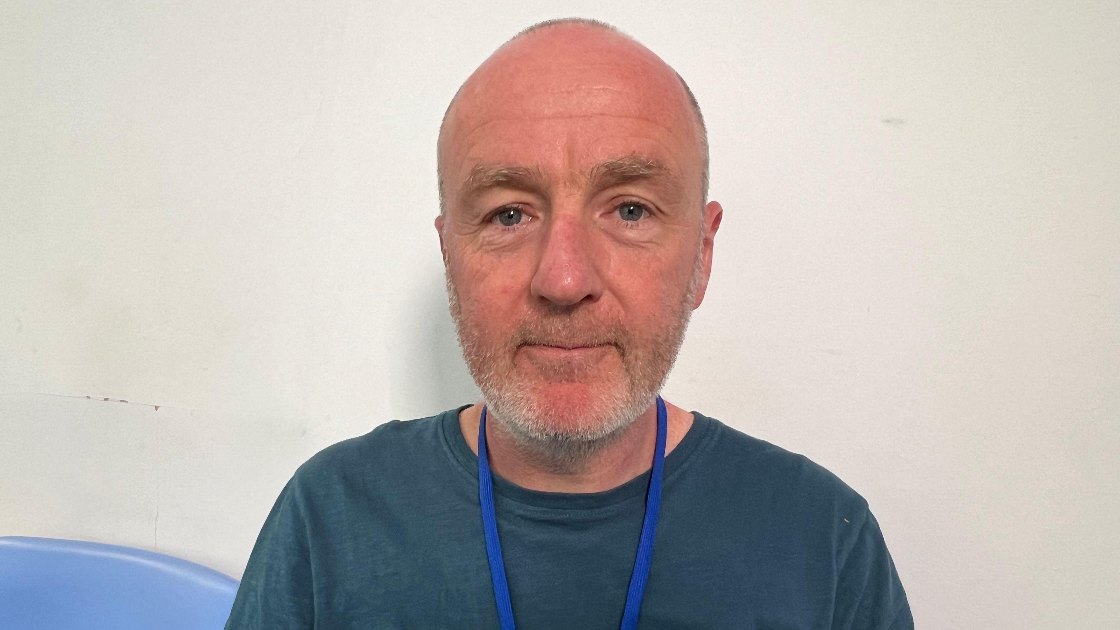 Matt Davidson wearing a grey-blue t-shirt and a blue lanyard. He is bald with a shaven beard and is looking at the camera, standing against a white wall. 