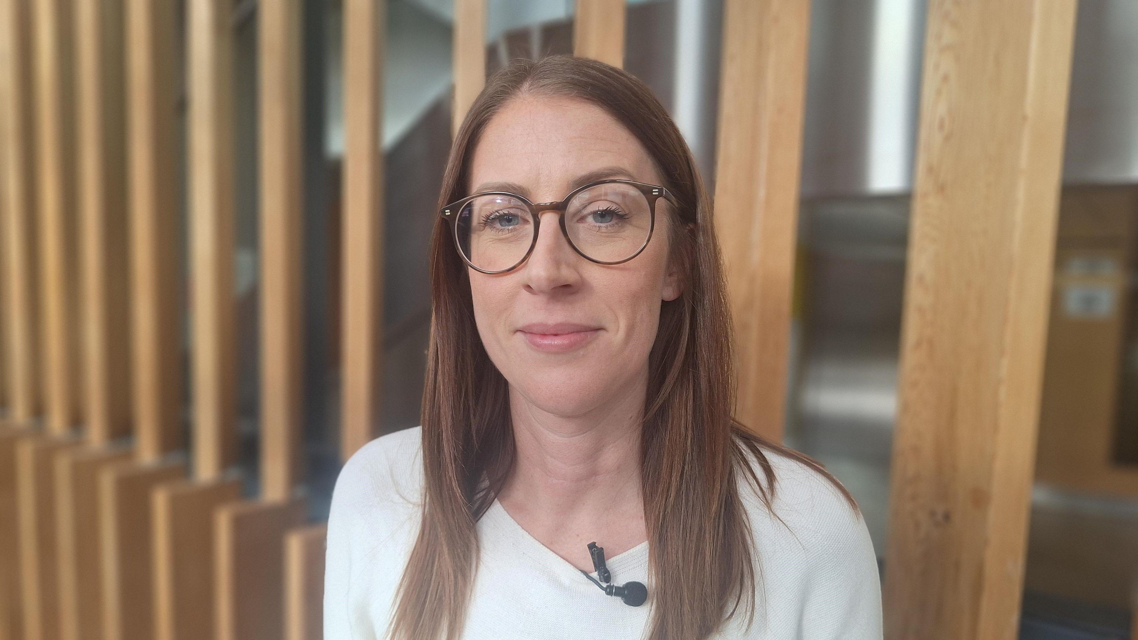 Sophie Legge is standing in front of a wall of wooden slats. She is wearing a white sweater and is looking into the camera with one eyebrow slightly raised. She has long brown hair and is wearing brown glasses.