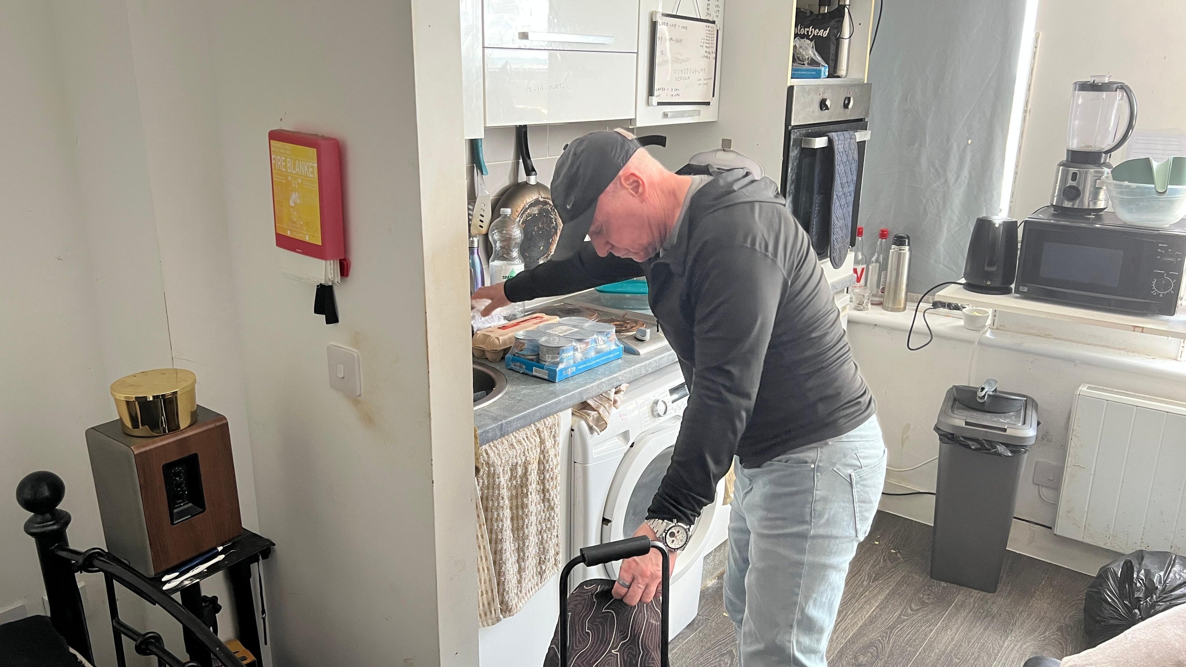 Glen Lane wearing a baseball cap, hoodie and light blue jeans unpacking his shopping in a small kitchen. In the foreground is the edge of the bed.