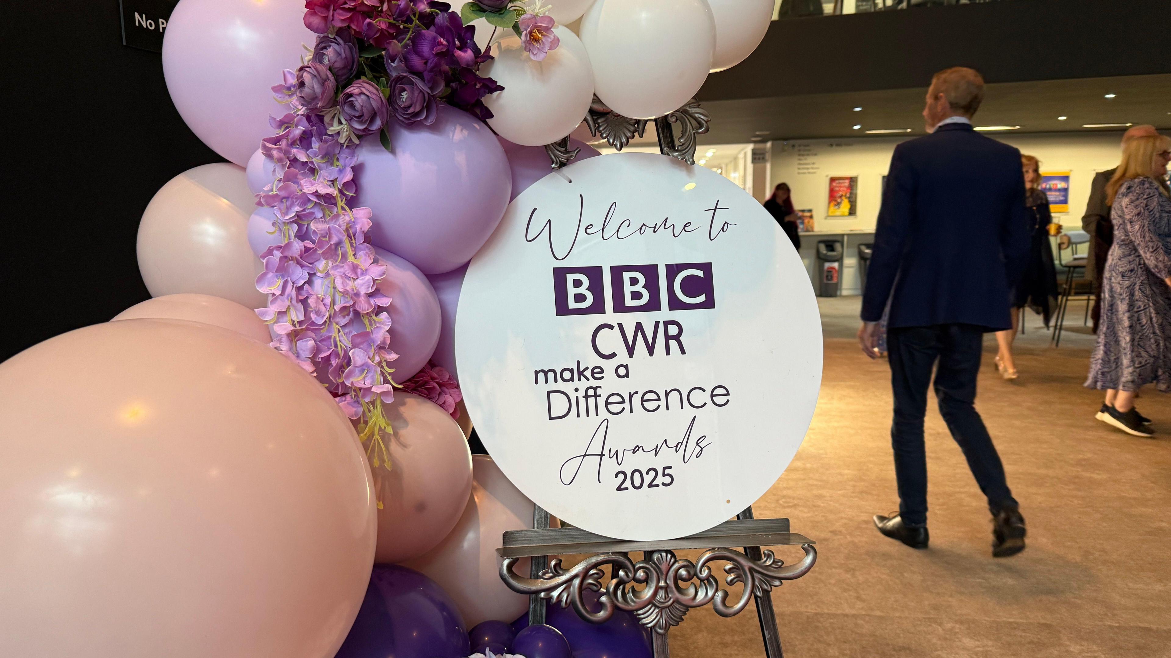 A white circular sign reads "Welcome to BBC CWR Make a Difference Awards 2025". There are pink and purple balloons around the sign with a man in a dark suit walking past the display.