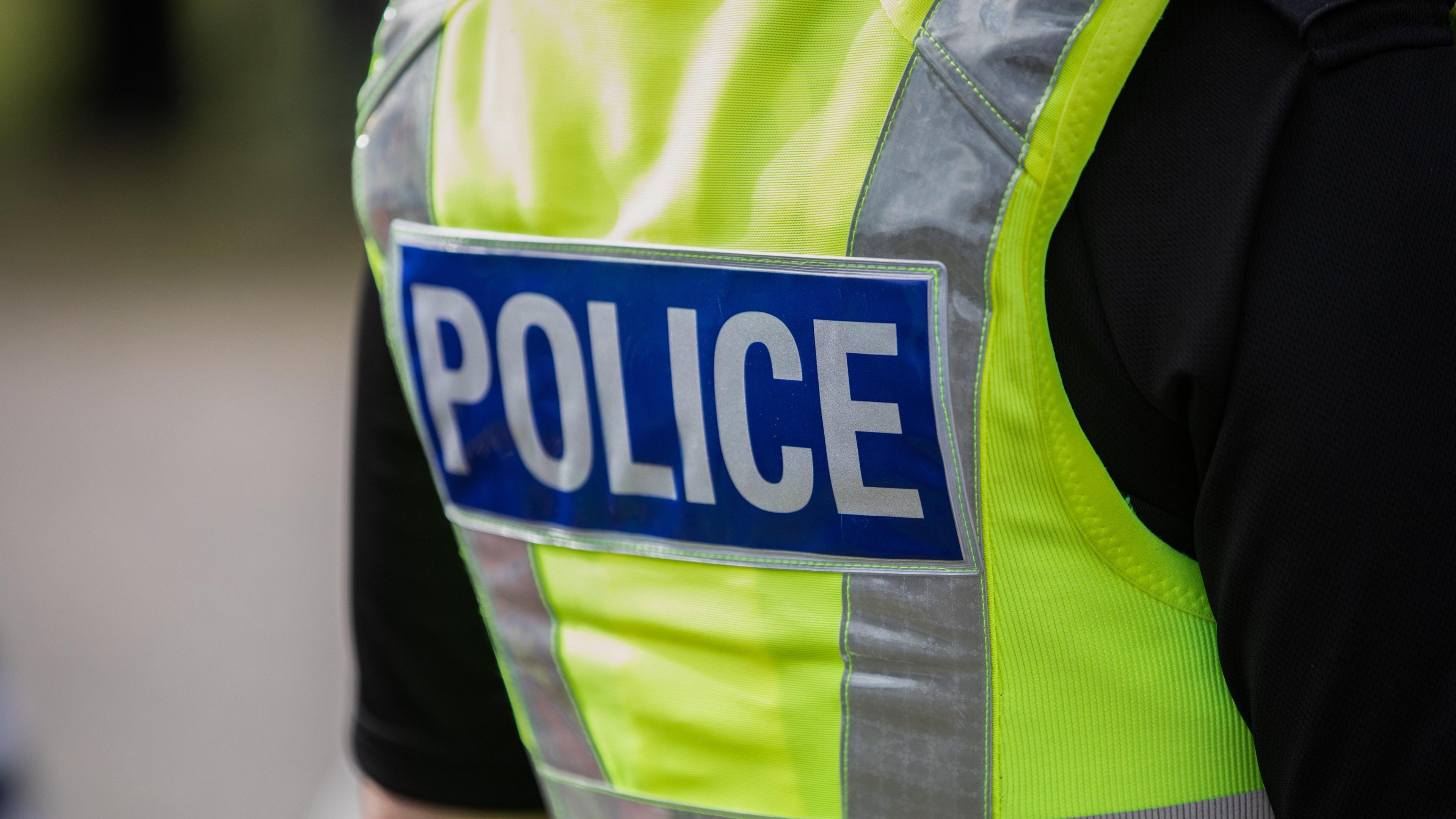 Image of the back of a high vis police vest. It is neon yellow with silver reflective stripes and a blue rectangle with police written on it.