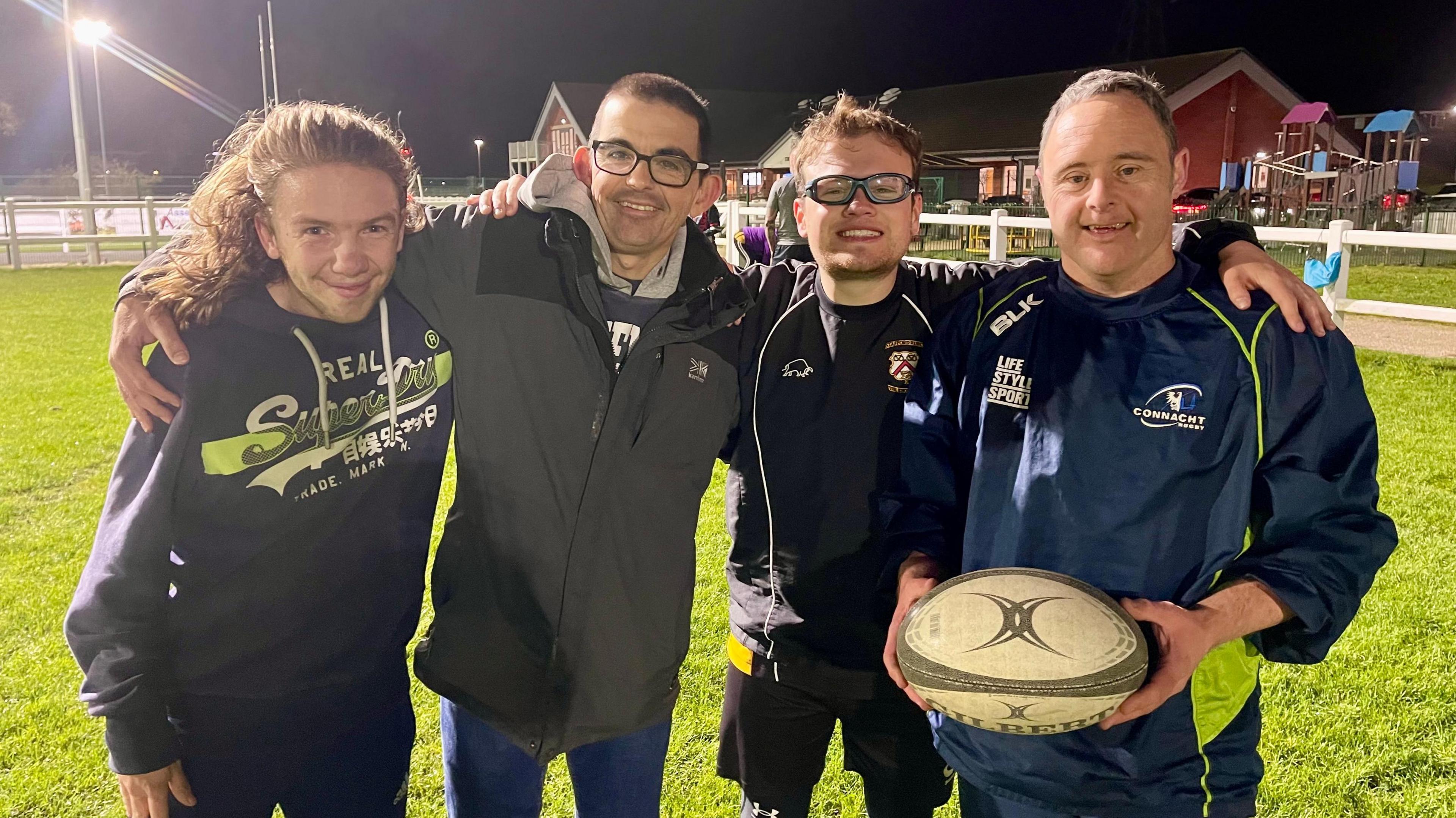 Four men embrace on a rugby pitch with the man on the right holding a rugby ball. The man on the left has long hair and is wearing a navy hoodie. Next to him is a man in a grey coat and glasses. Next to him is a man in glasses, a blue tracksuit top and shirt. To the right is a man in full navy tracksuit. They are all smiling under the floodlights.