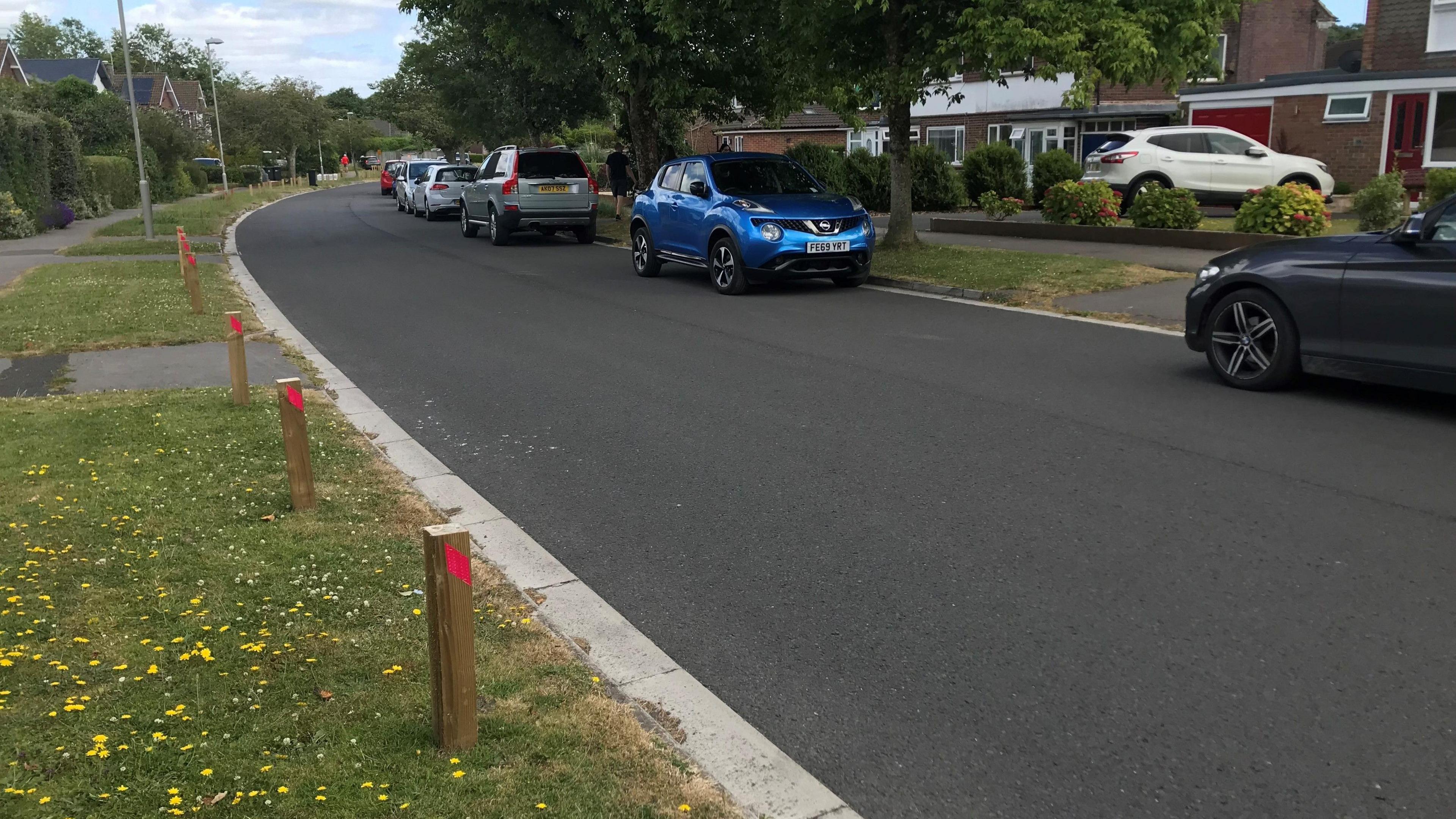 A residential street lined with grass verges and trees. There are cars parked on one side and a line of wooden posts in the grass on opposite verge