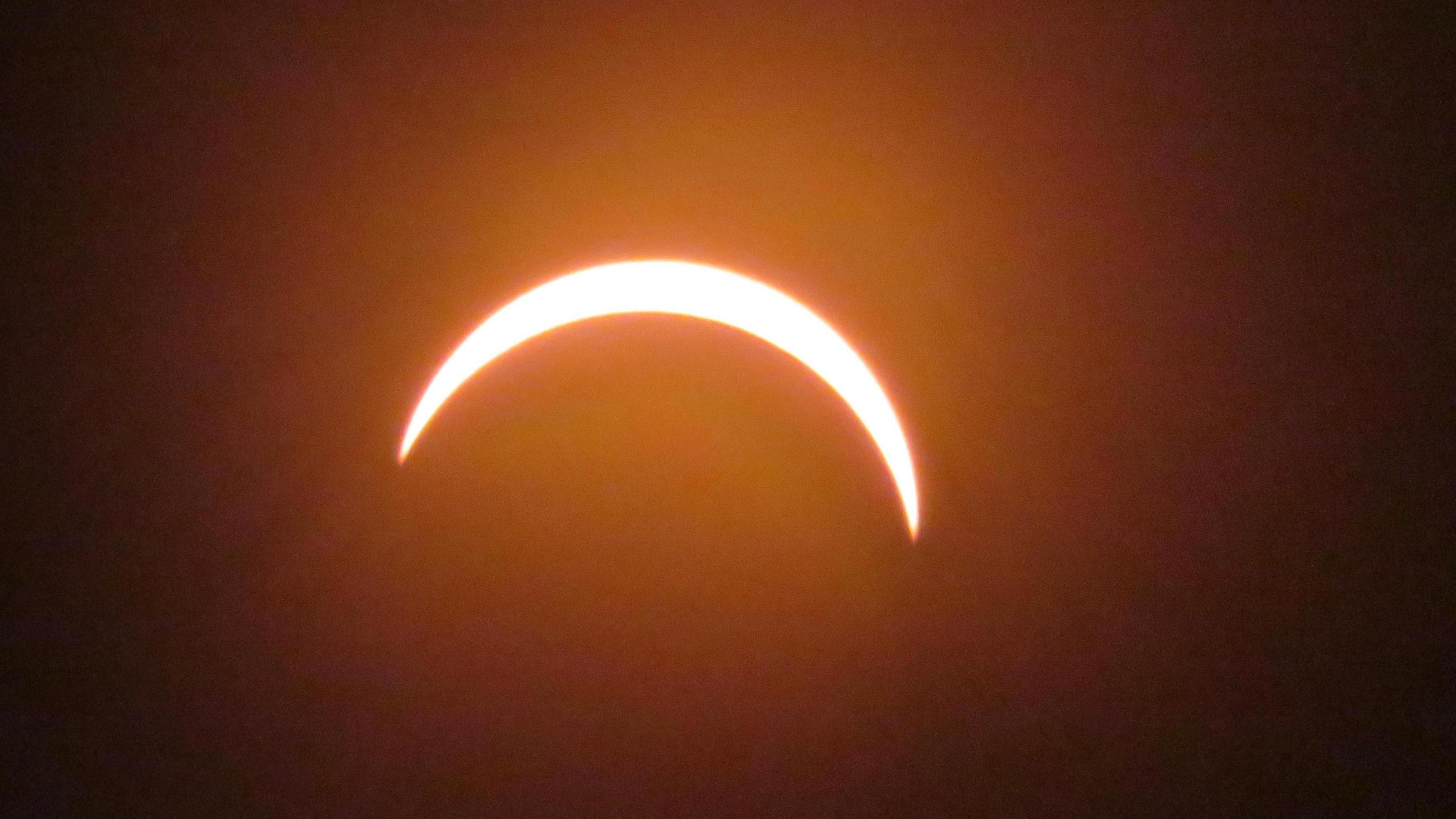 A close-up photograph of the partially eclipsed Sun in Lamberhurst, Kent. The Moon covers most of the Sun, leaving a bright crescent-shaped arc of light against an orange-tinted sky.
