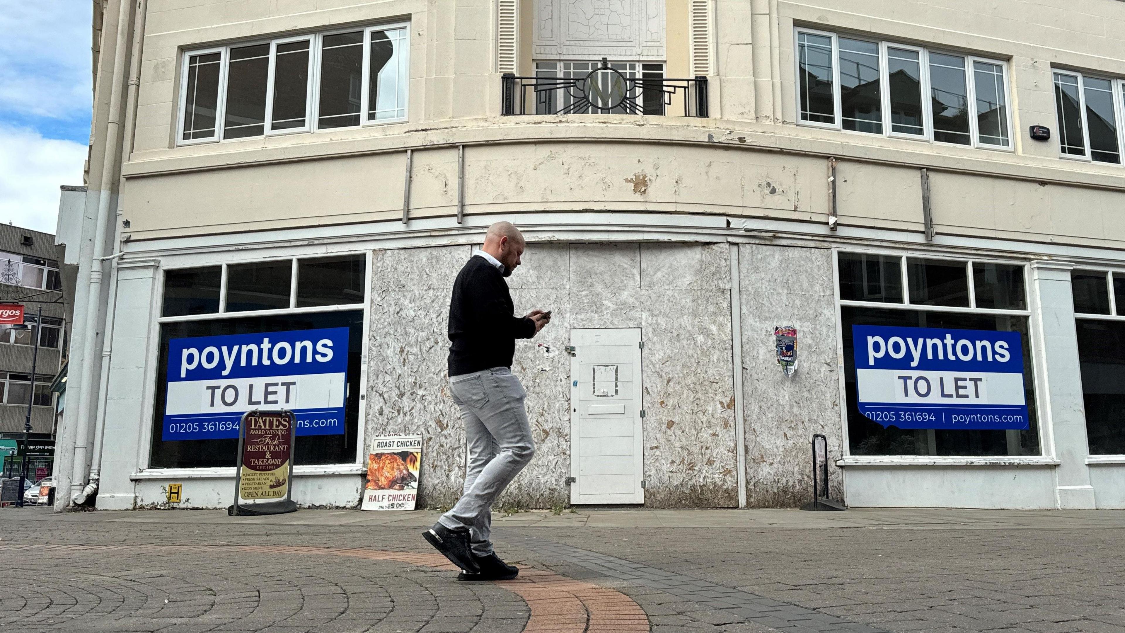 A man walks in front of an empty shop. The entrance is boarded up and  large 