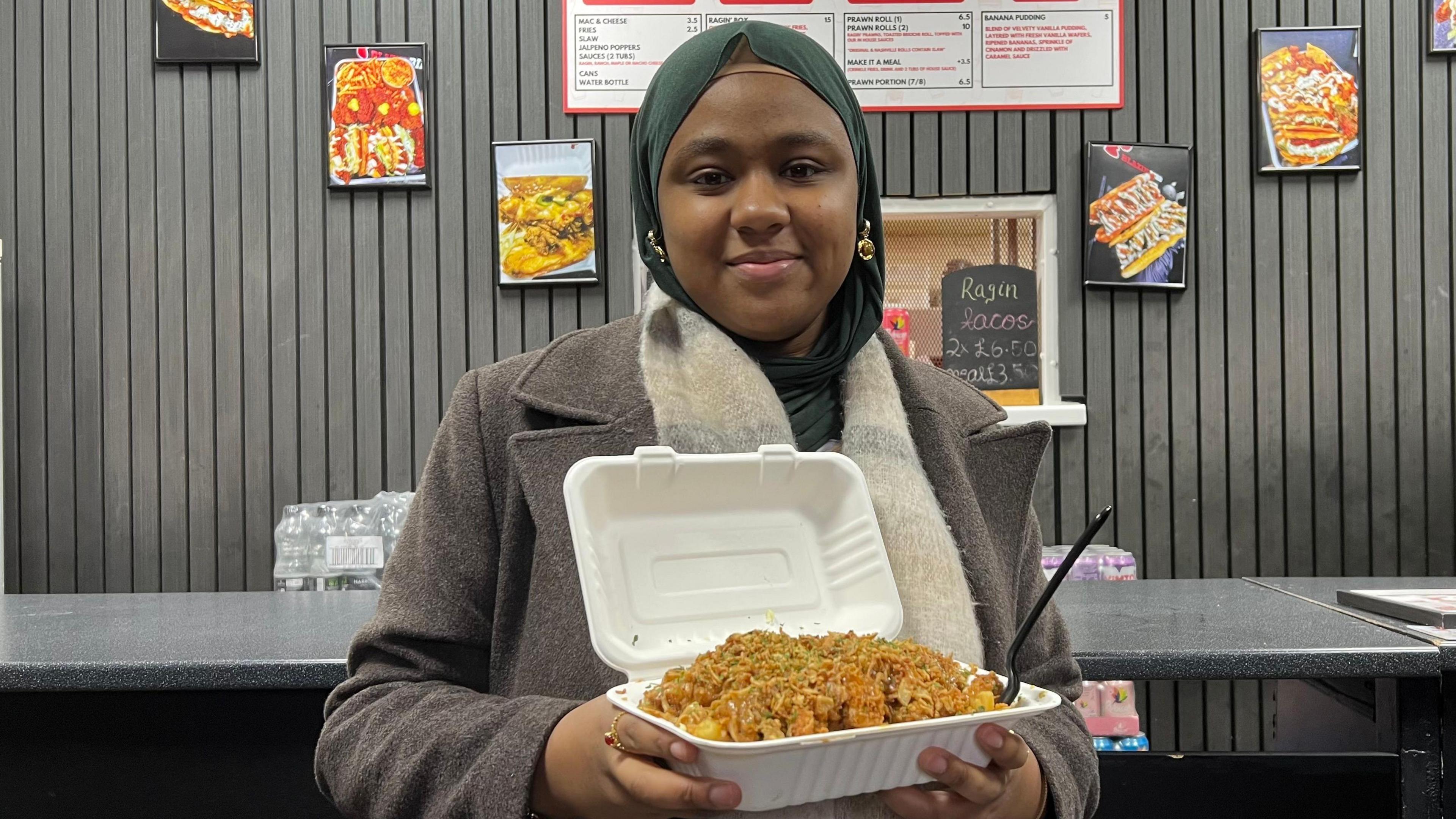 Sumeja ka veshur një pallto të gjatë ngjyrë bezhë-gri dhe një hixhab ose shami jeshile. Ajo mban një kuti të bardhë të hapur me ushqime për të marrë me vete, të mbushur me pulë të skuqur të erëzuar brenda një dyqani ushqimi të shpejtë, me tabela menuje dhe foto ushqimesh të shfaqura në murin me panele të errëta pas banakut.