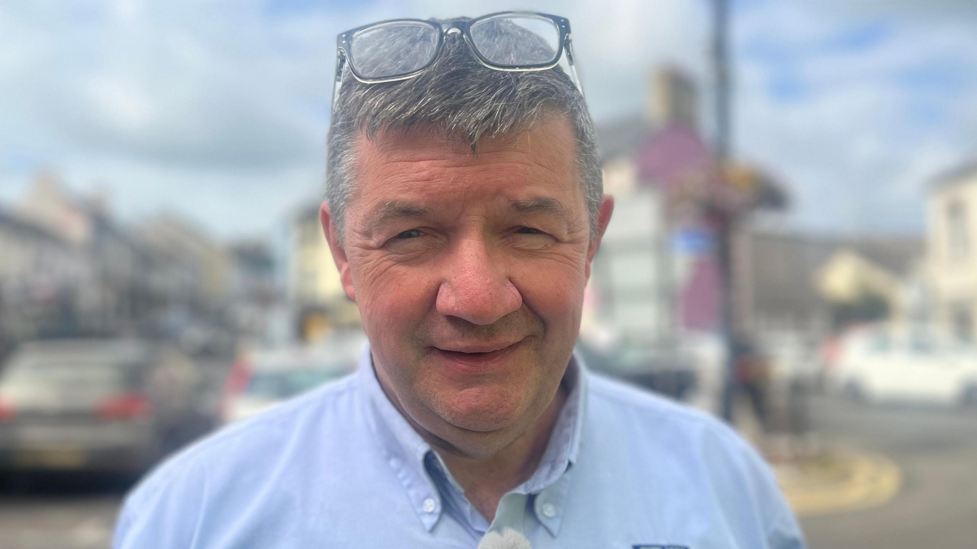 Paul Cochrane stands on the street in Ballycastle. He wears a light blue shirt and has grey hair. His glasses are on his head.