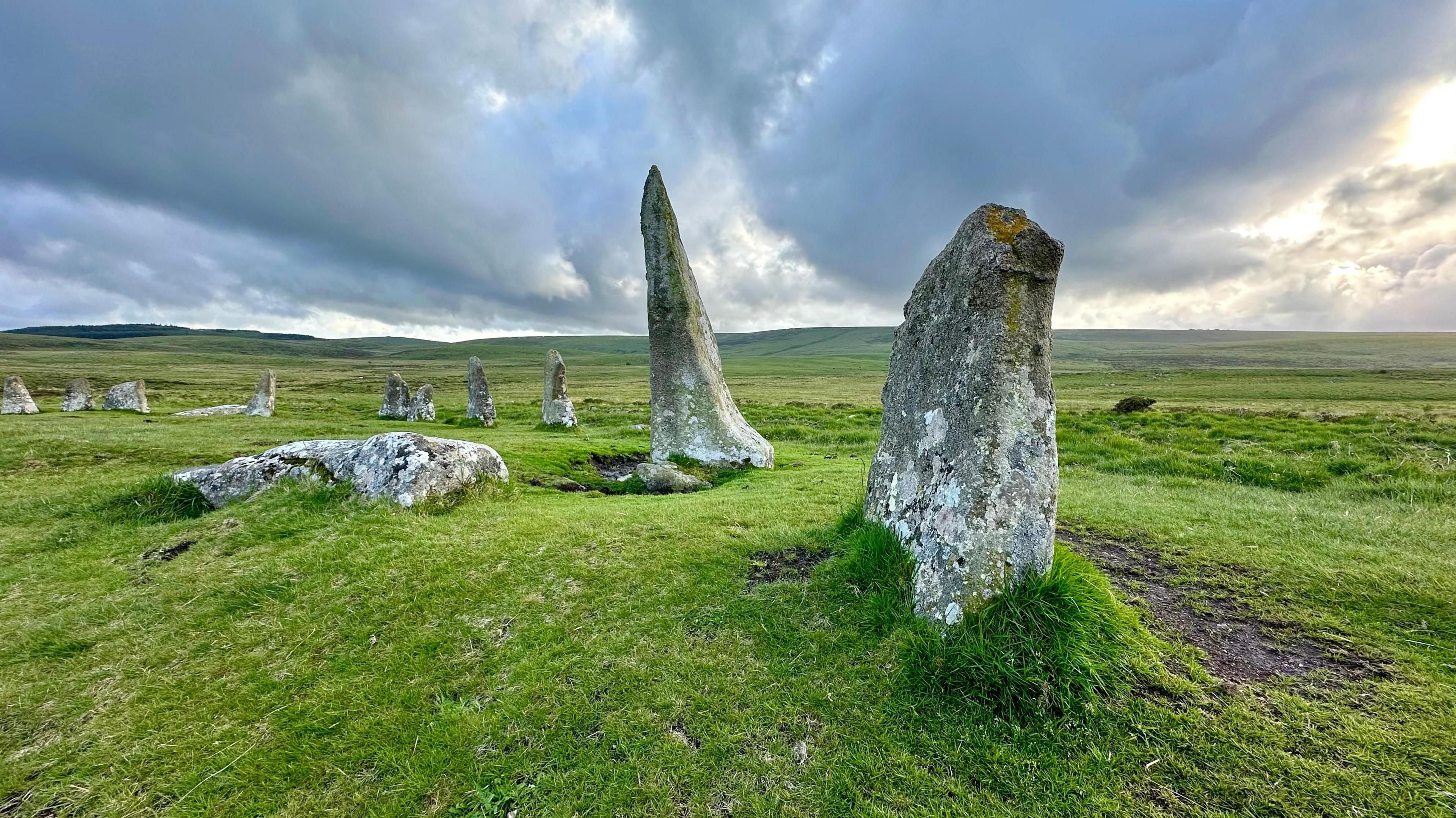 Scorhill stone circle on Dartmoor. Overcast weather conditions above the stones, which are pointy. One stone is lying flat on the grass.