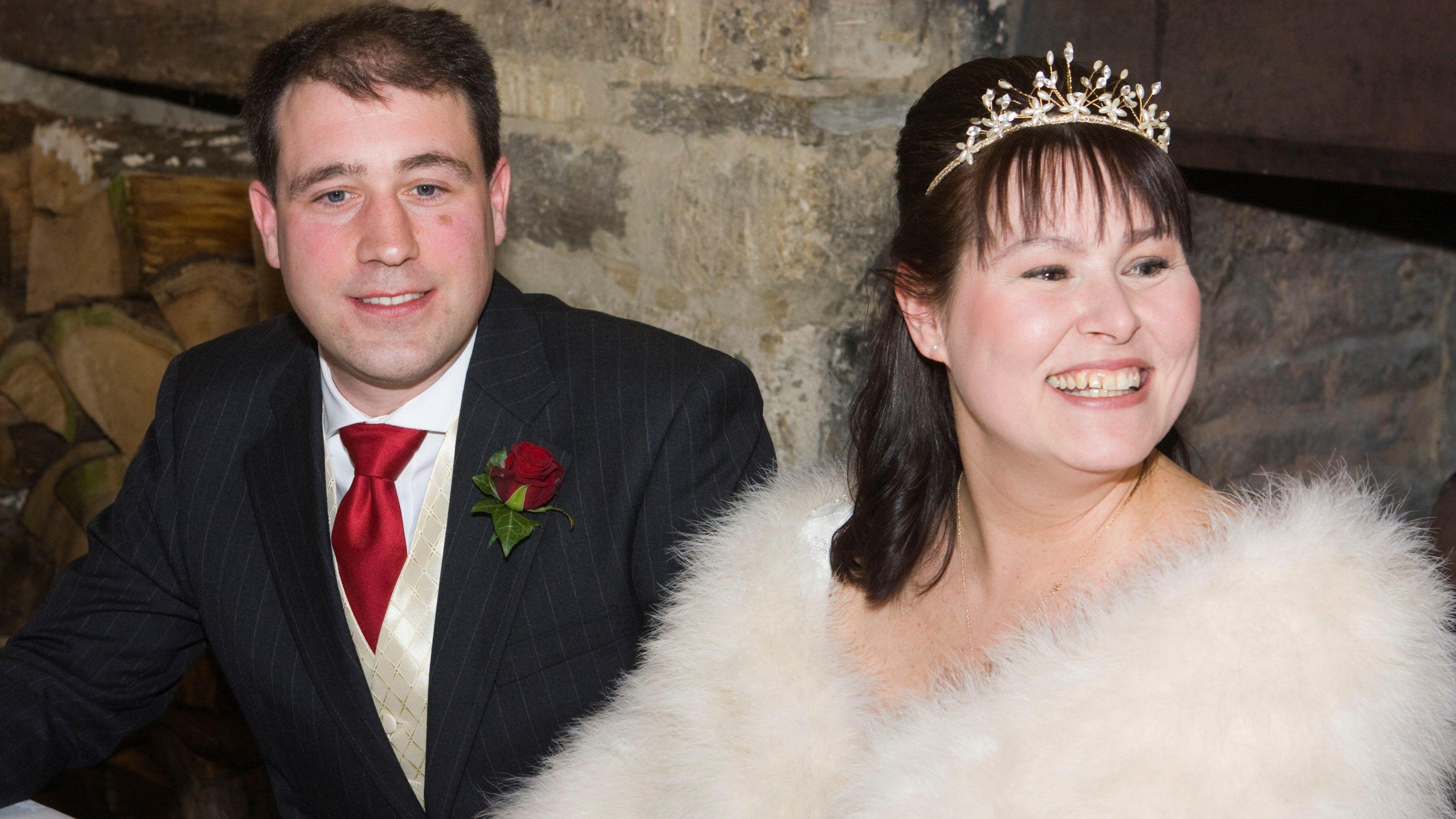 A picture of Jonny (left) and Anya on their wedding day. Jonny wears a three-piece suit with a red tie and a red rose buttonhole, while Anya wears a tiara and a white fur wrap. 