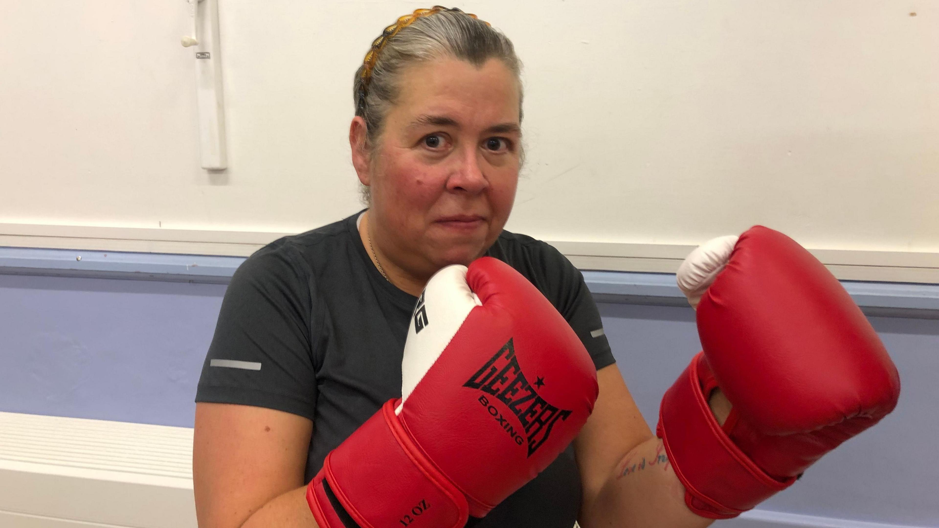 Linda Smith, 53, has blond-coloured hair tightly swept back. She is wearing a grey t-shirt and a gold necklace. She has red and white boxing gloves with black writing on them saying 'Geezers boxing'.