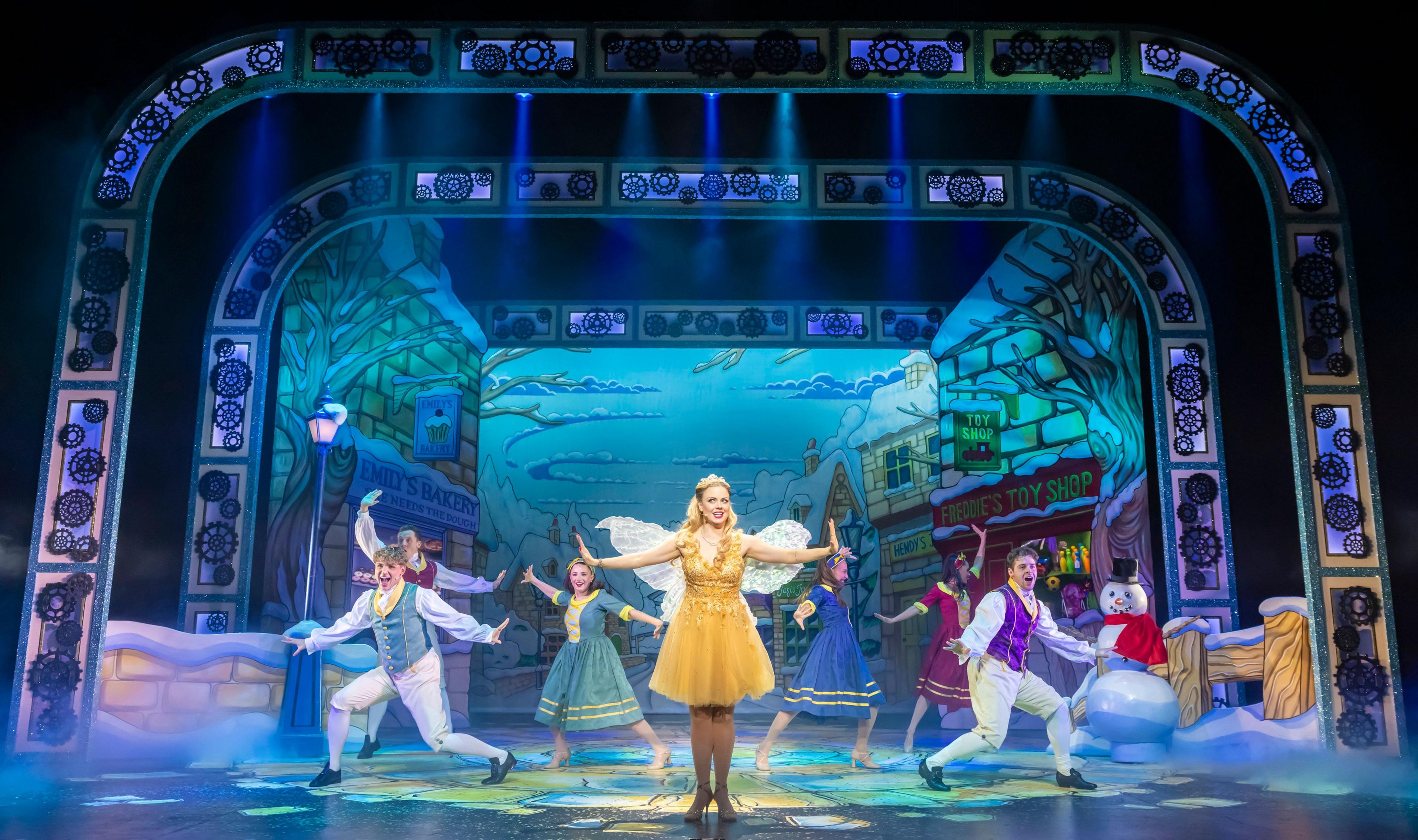 Joanne Clifton, wearing a gold Fairy Godmother outfit, in a pantomime, with six dancers, with their arms out, around her. There is also a snowman prop to the right. There is a snow scene behind them and lights on the stage.