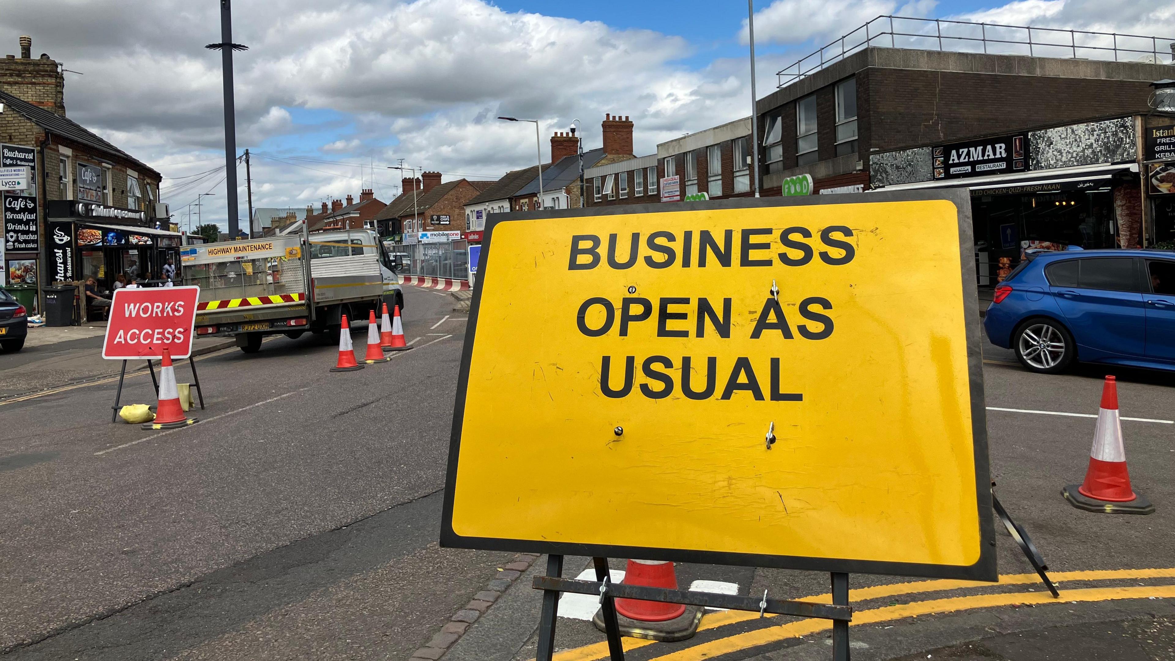 A yellow road sign which reads 'Business Open as Usual' stands on a road. Behind it is a road with cones, a maintenance vehicle and more road signs.