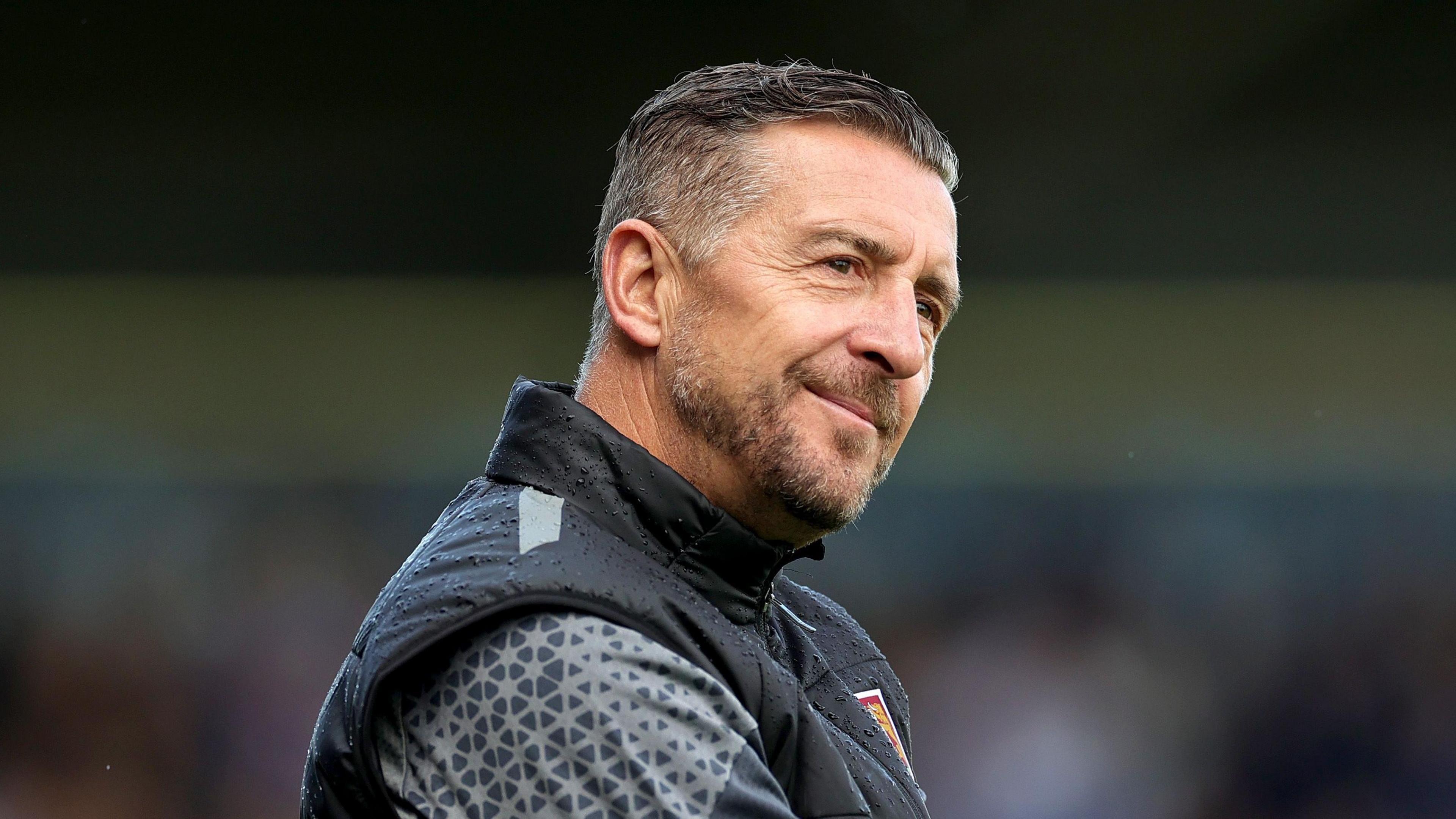 Jon Brady wearing a black Northampton Town tracksuit. He has gelled dark hair and is looking into the distance with his head tilted sideways. 