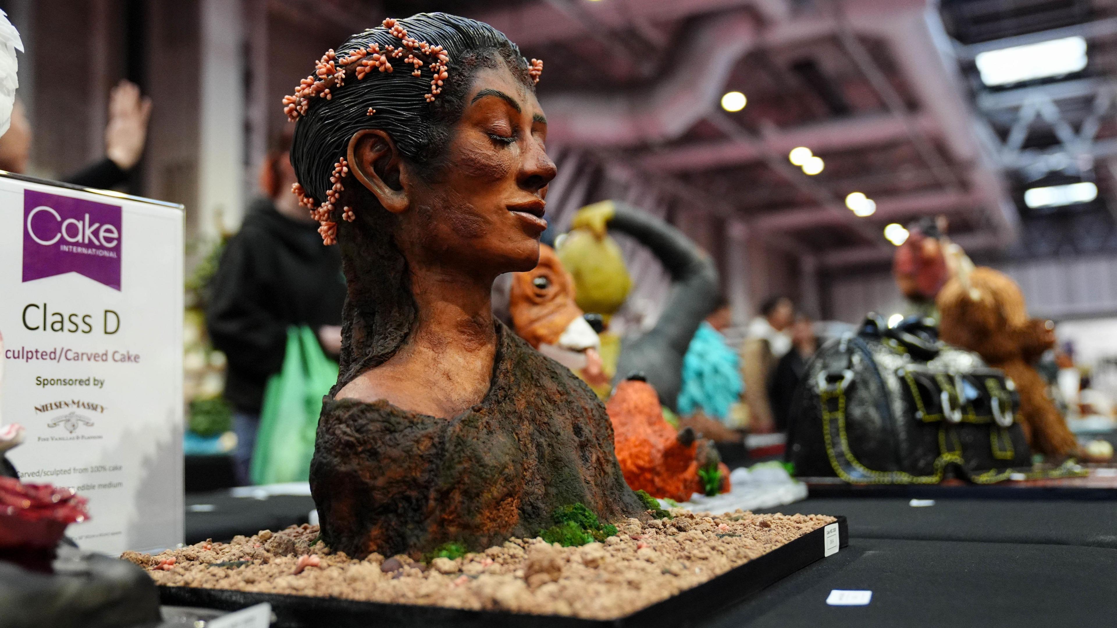 A cake display depicting the head, shoulders and upper torso of a woman with long black hair and a dark top which is dropping off her right shoulder. She has her eyes closed and is smiling. The display has been placed on a black table in an exhibition room.