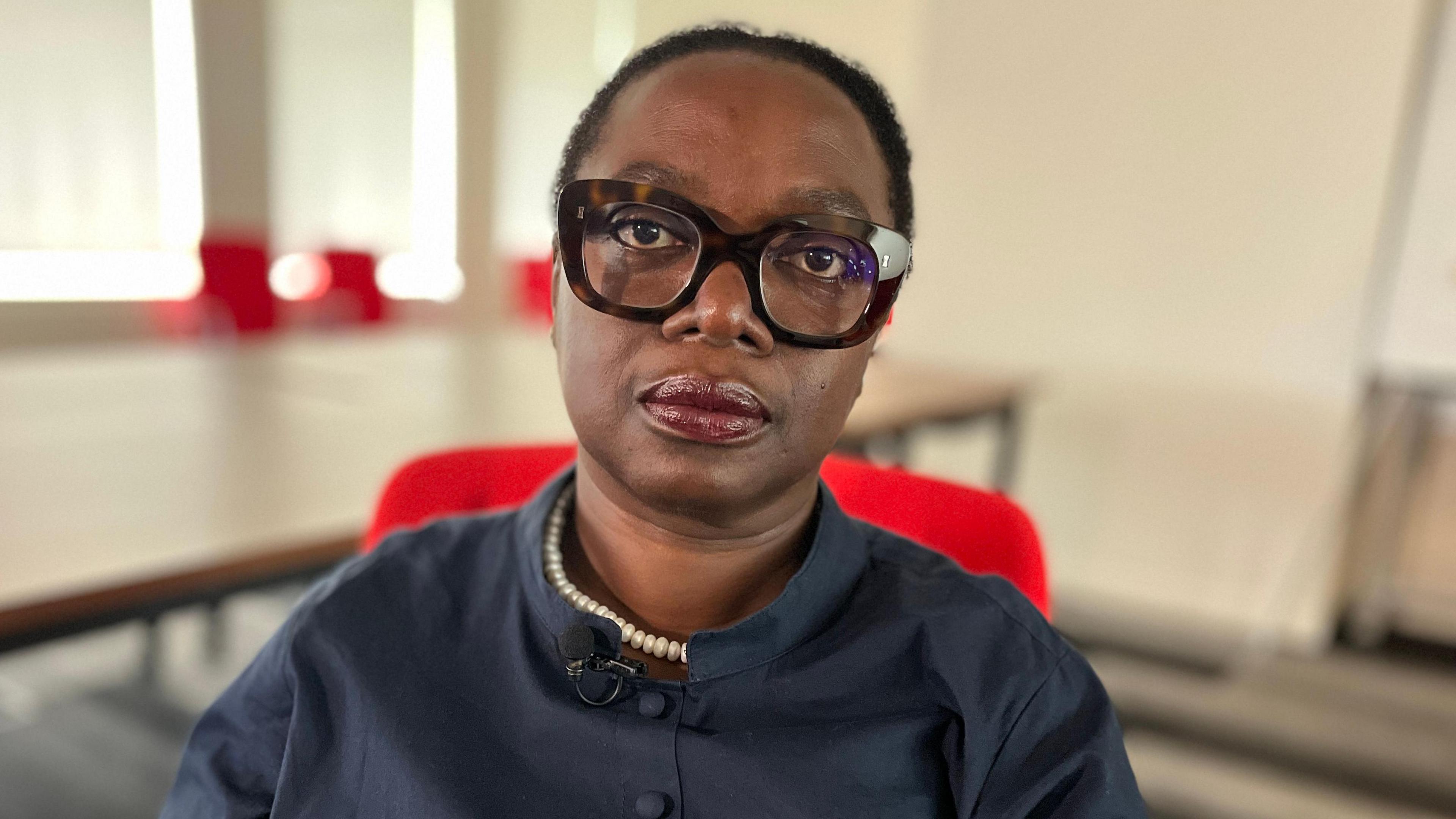A woman sitting in a red chair within a room looks at the camera. The background behind her is blurred. She has dark, short hair, and is wearing large glasses along with a navy blue blouse and pearl-style necklace.