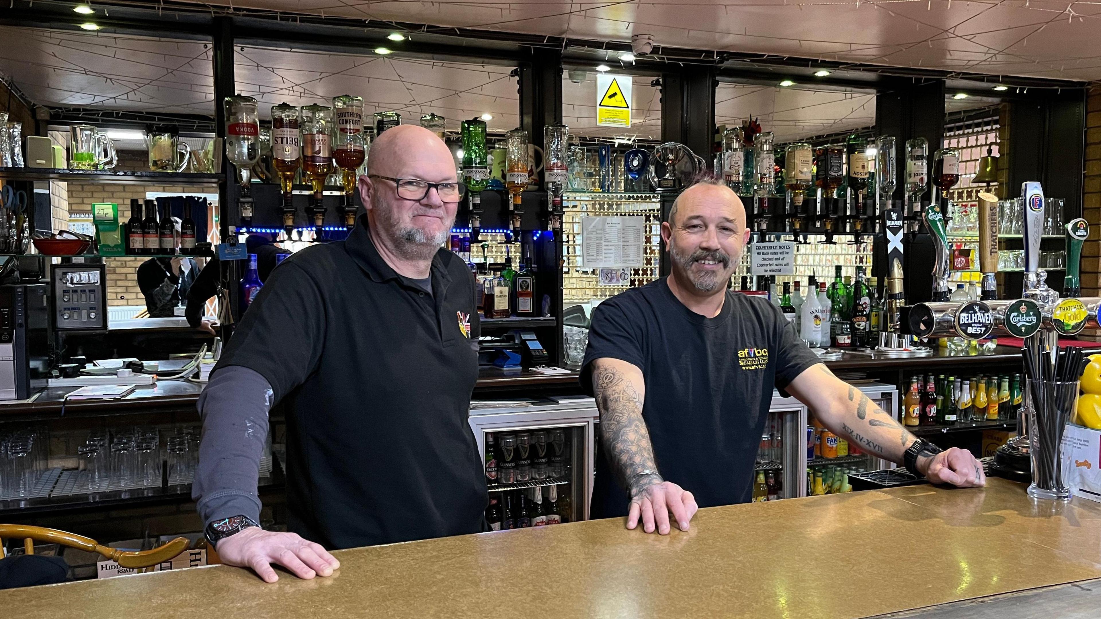 Sean Howlett and Carl Higgs are stool behind a bar together. They have their hands on the counter and are smiling at the camera. Sean is bald and has grey facial hair. He wears square framed glasses, a black top and a long sleeved grey top underneath. Behind them are bottles of alcohol and draught beer taps are on the bar.