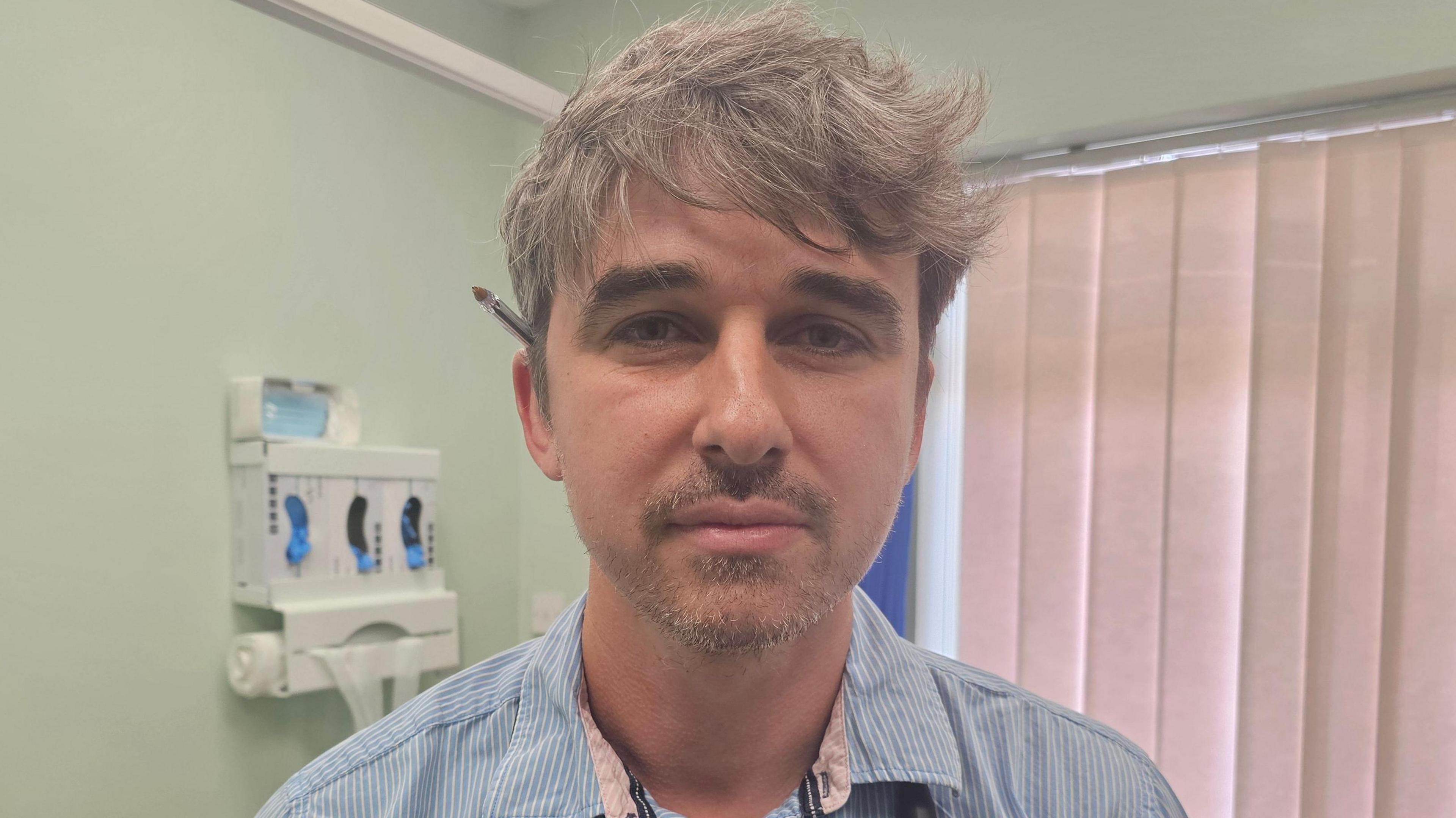 Dr Matthew Jones with short brown hair and a slightly greying brown beard. He has a biro ball point pen tucked behind his ear and is looking directly at the camera. He has a blue shirt with small white stripes and the background is lime green walls, with a dispenser for glove and waste bags. There is also a window covered by cream blinds.