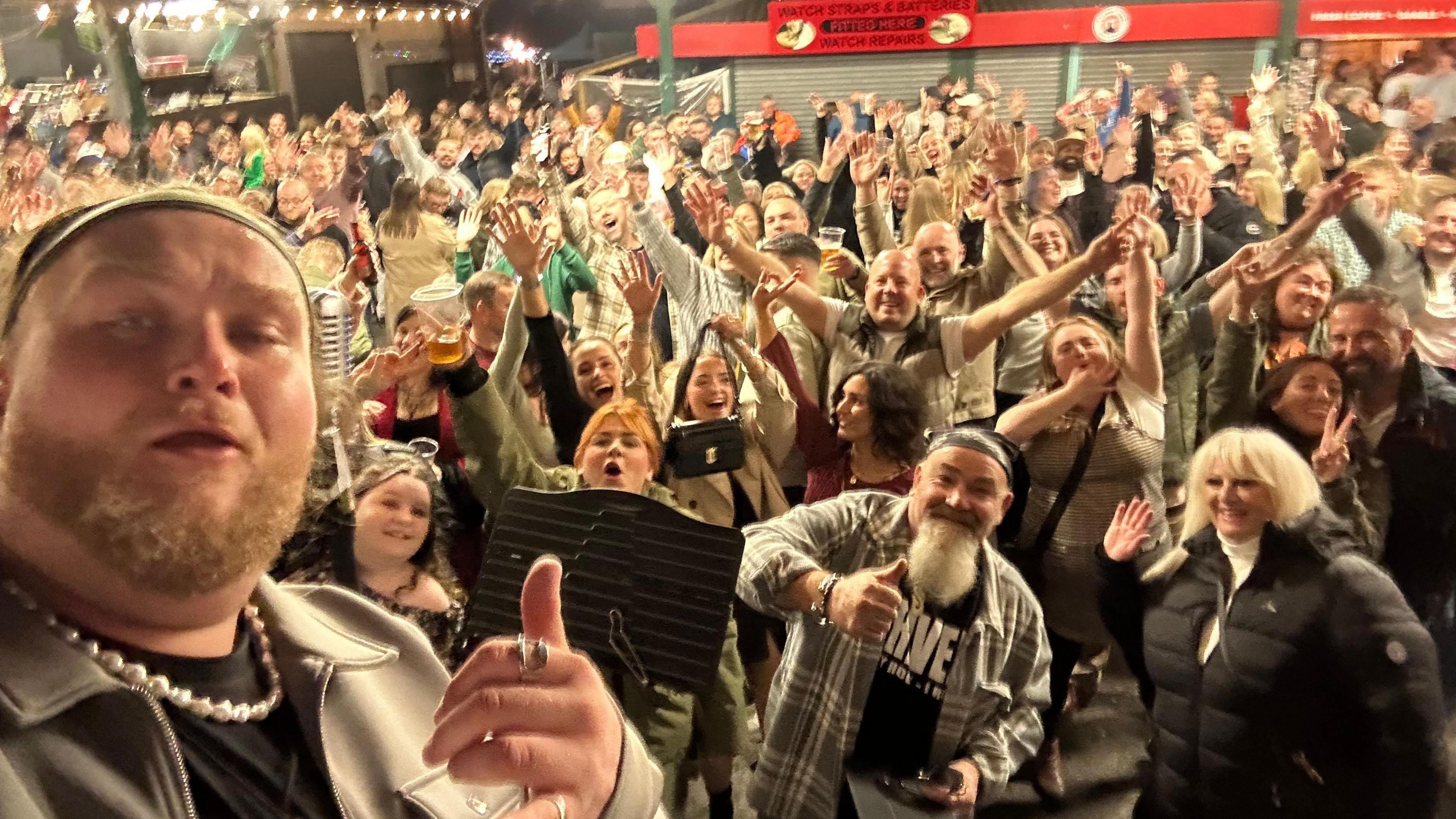 Callum Doignie is taking a selfie from on stage. He has brown facial hair and is wearing a hat, with his hand up. Behind him are hundreds of people in a crowd, waving at the camera.