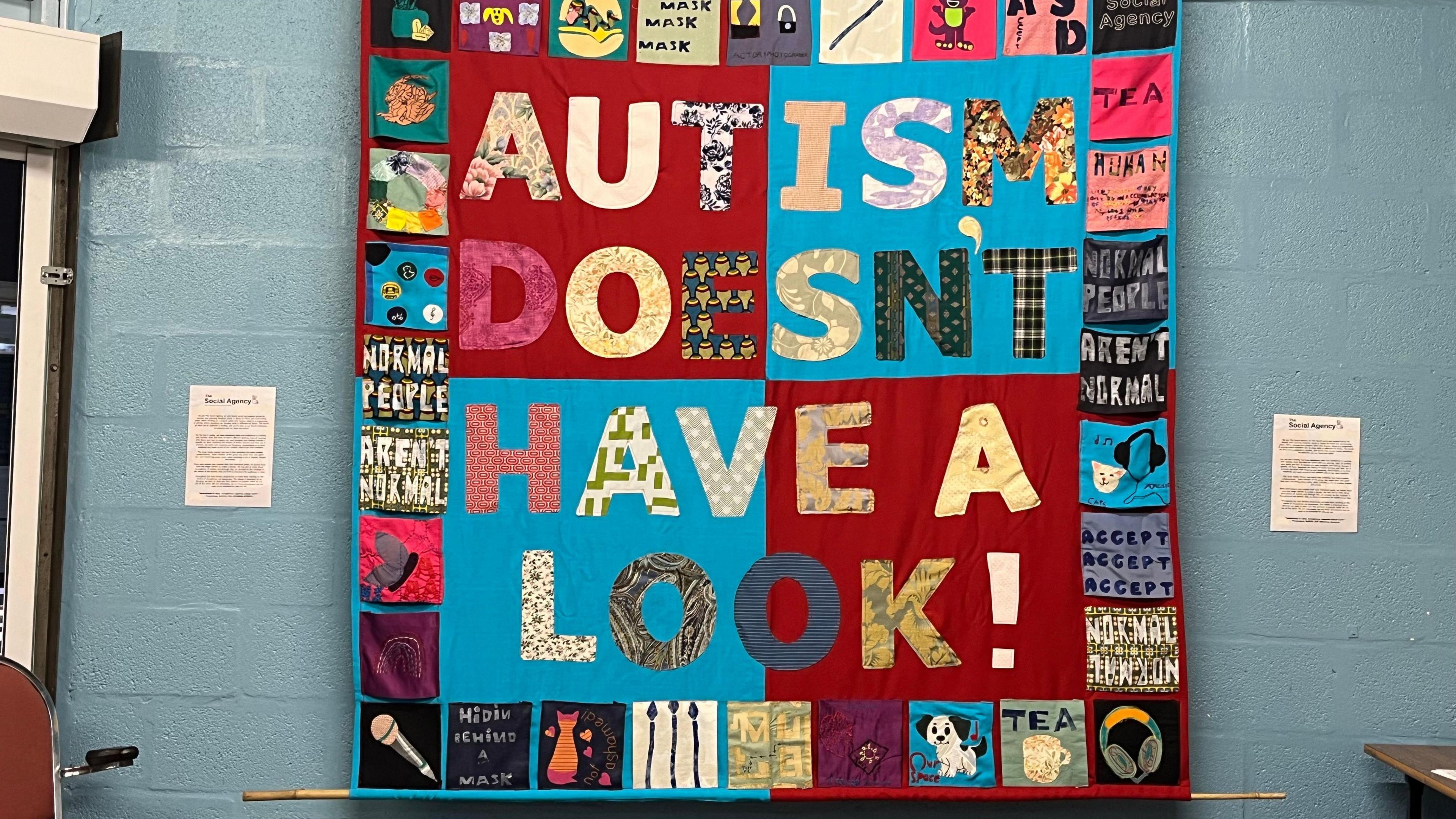 A banner is displayed on a wall created out of textiles with red and blue panels and the words Autism Doesn't Have a Look made out of different fabrics. There are square panels around the edge of the banner, some with pictures of objects, and others with words including "normal people aren't normal", "hidden behind a mask", "accept, accept, accept" and "tea".