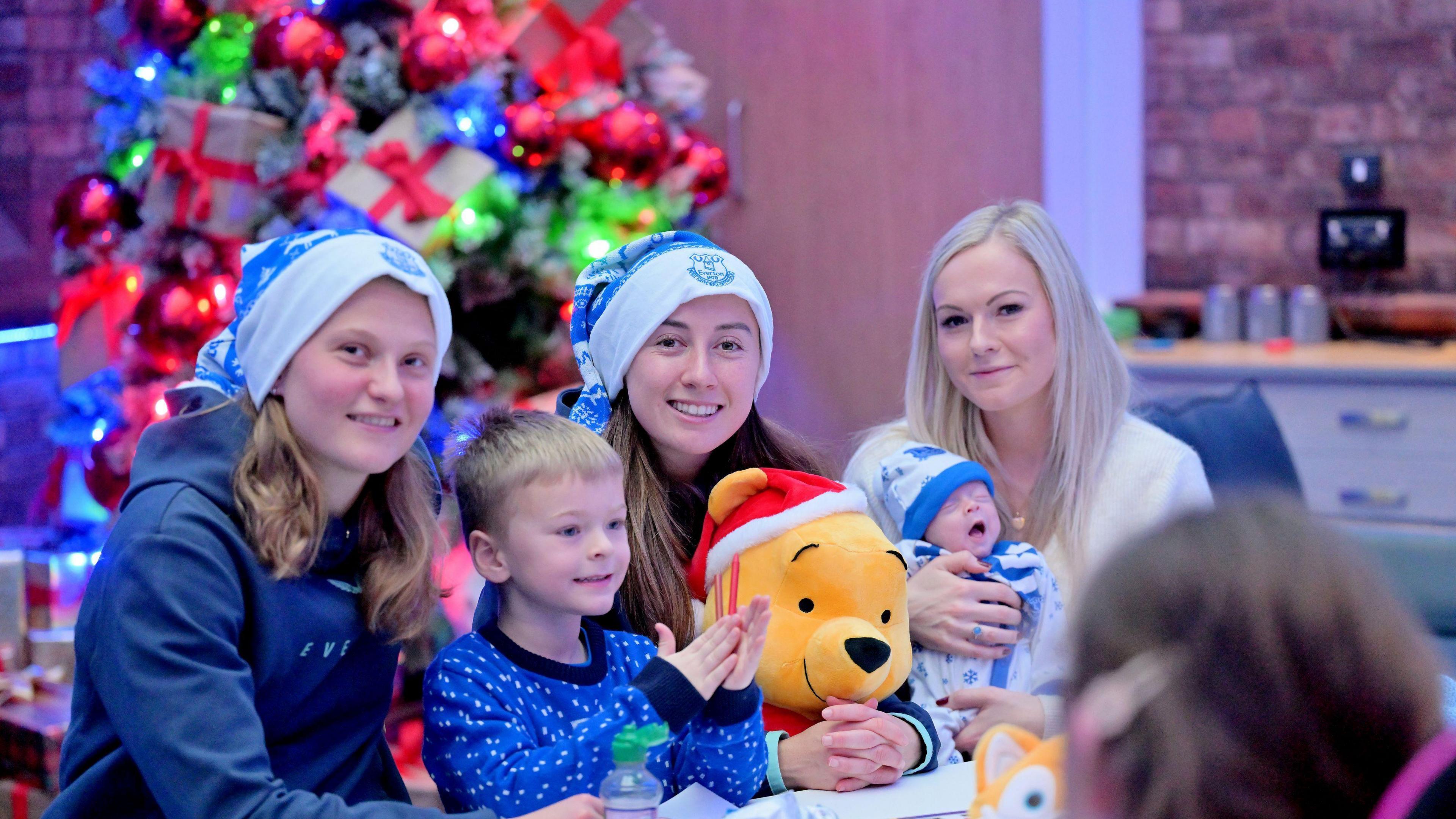 Elise Stenevik and Claire Wheeler, wear blue Christmas hats, holding a Winnie the Pooh toy, with one boy, and a mum and her baby