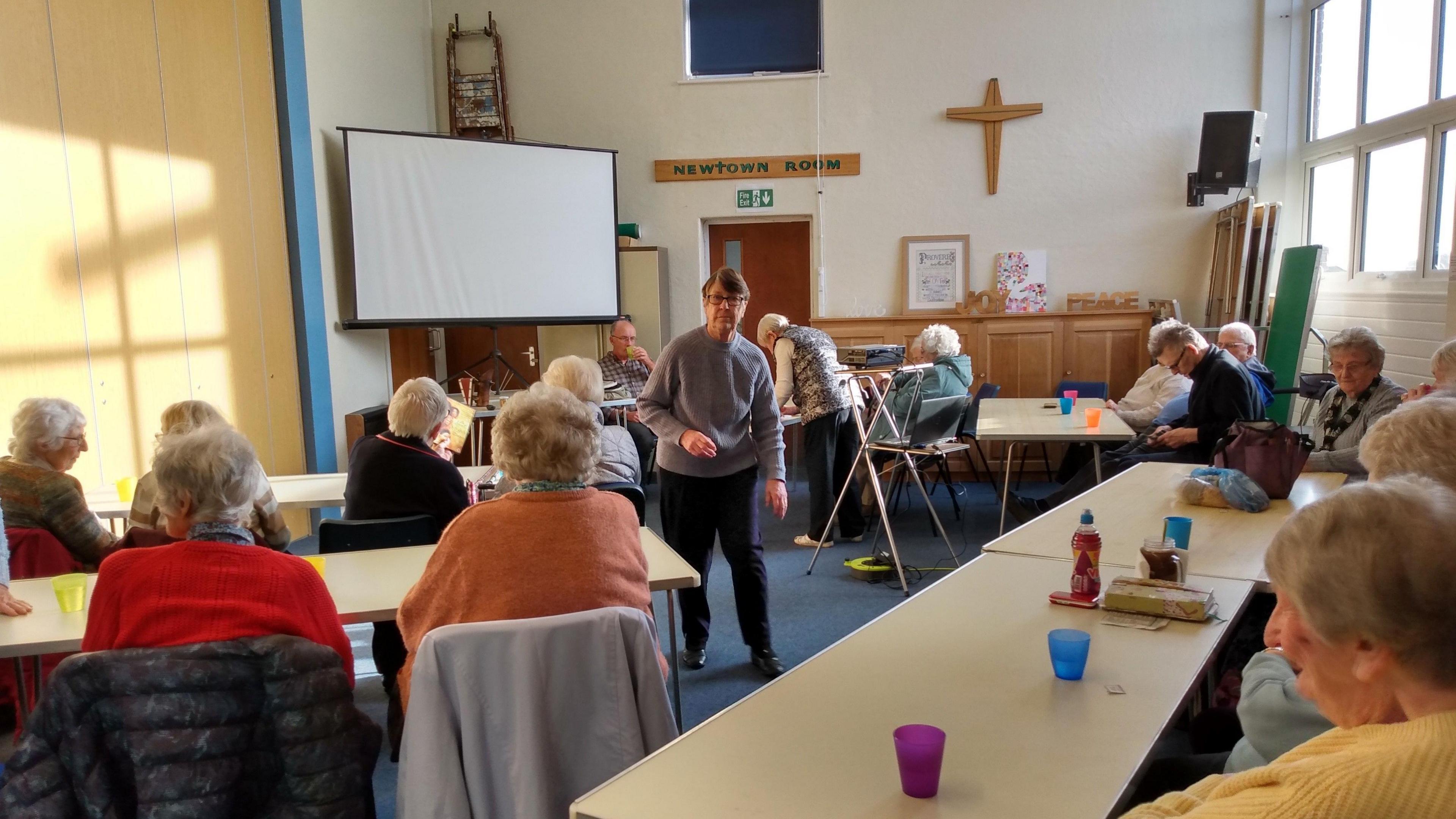 A room full of people with one lady in the middle who is leading the club. They are sat at desks with a large screen on the wall in the background and a projector on a tripod in the centre of the room.